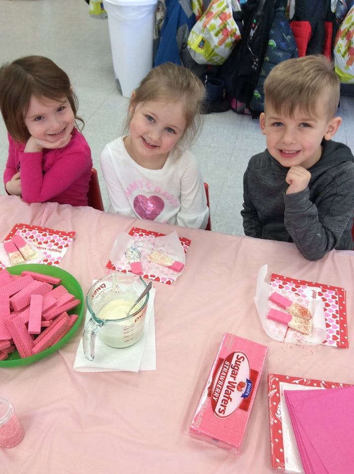 Three children sitting at a table with pink wafers and a measuring cup