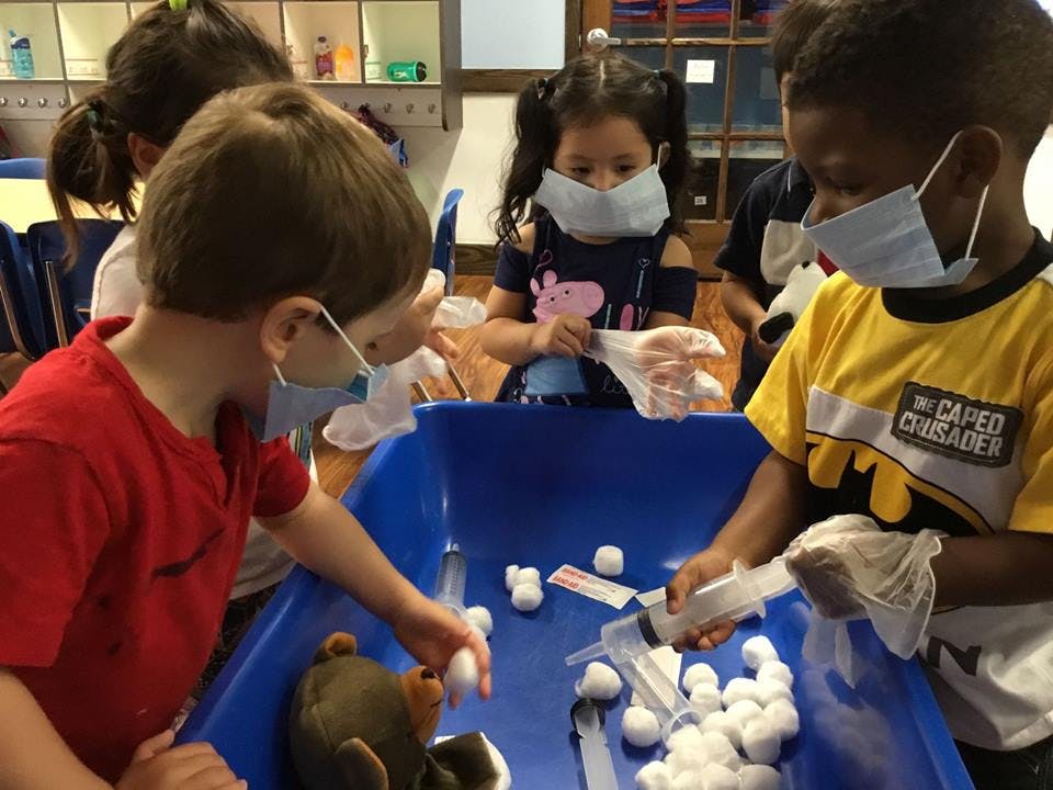Children in masks playing with cotton balls and syringes in a blue bin