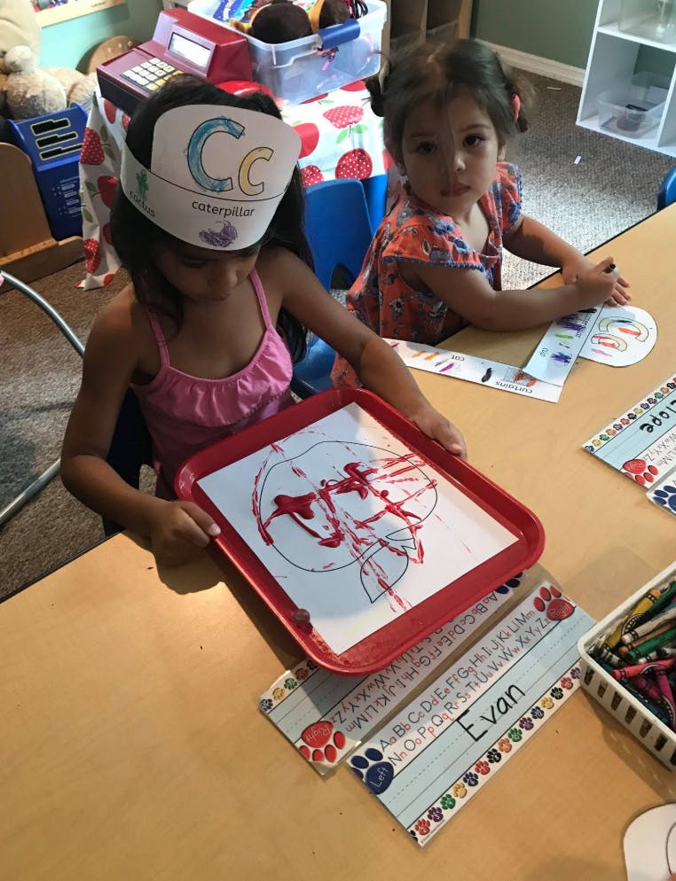 Two children at a table, one holding a tray with red paint on paper