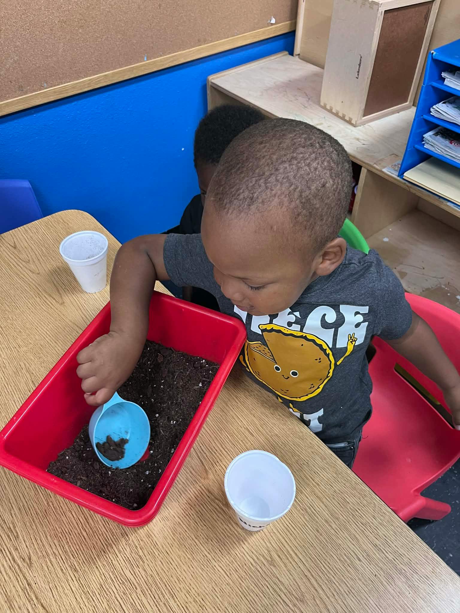 Child scooping dirt from a red tray with a blue scoop