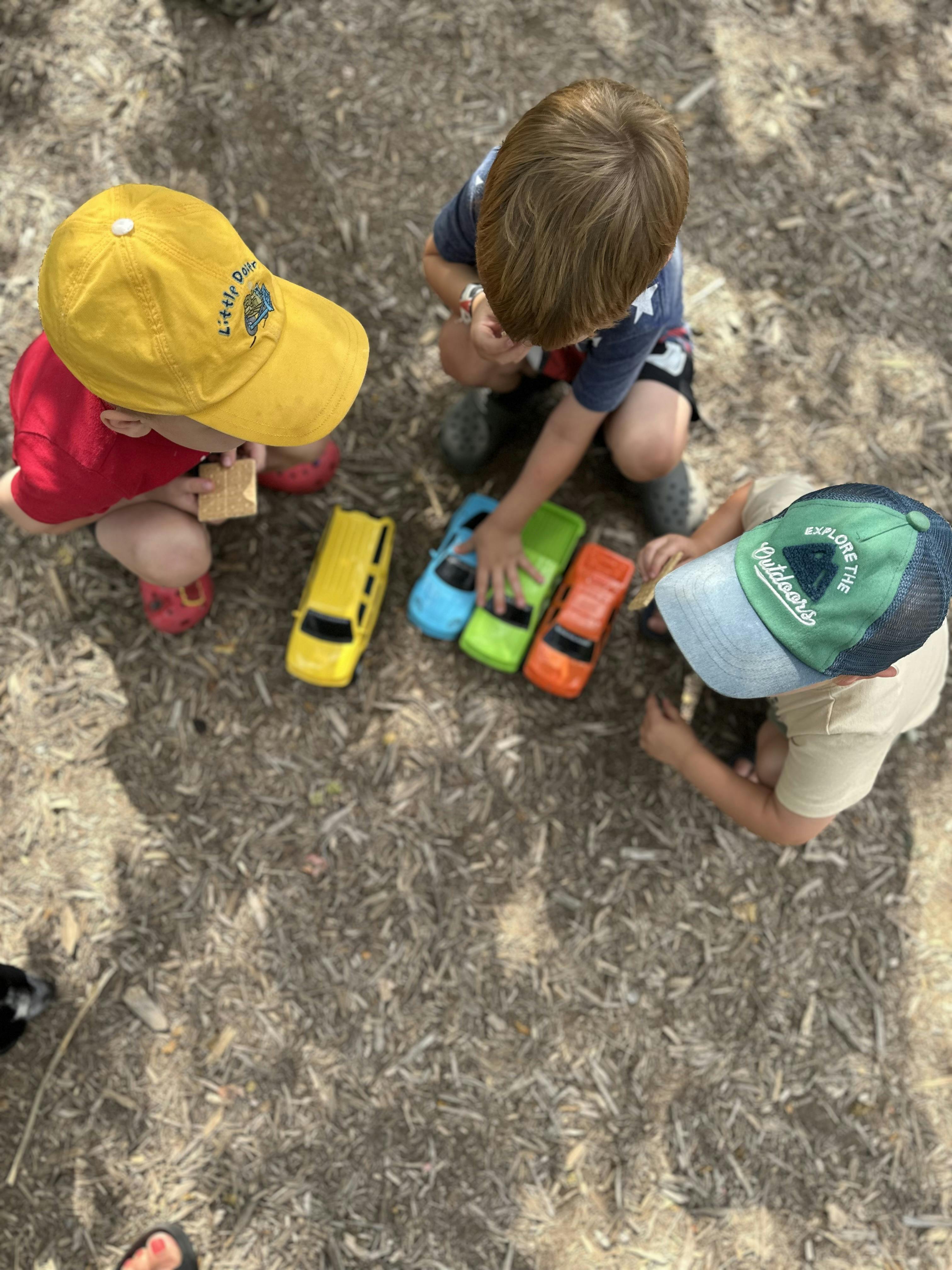 Three children playing with toy cars on wood chips
