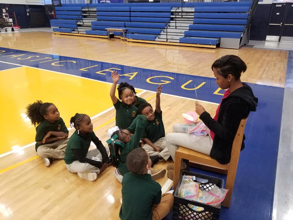 Adult and children sitting on a gym floor, some children raising hands