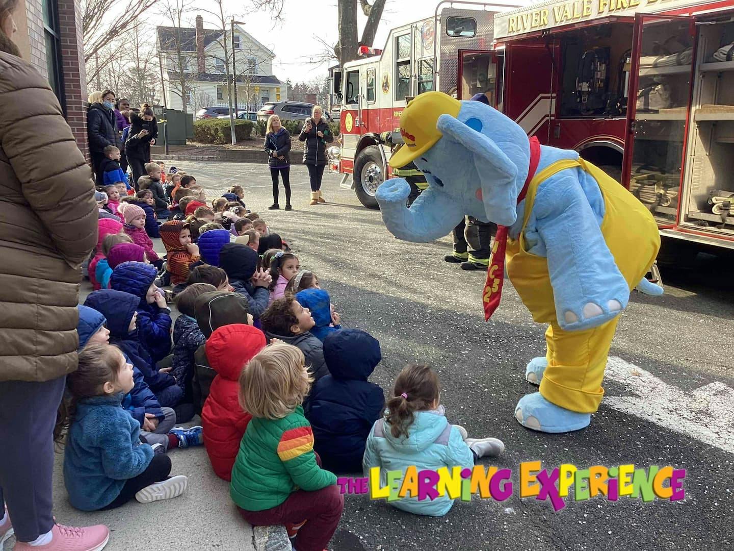 Children sitting on the ground looking at a person in an elephant costume