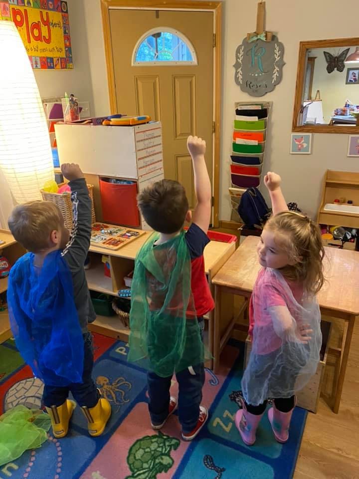 Three children in capes with arms raised in a classroom