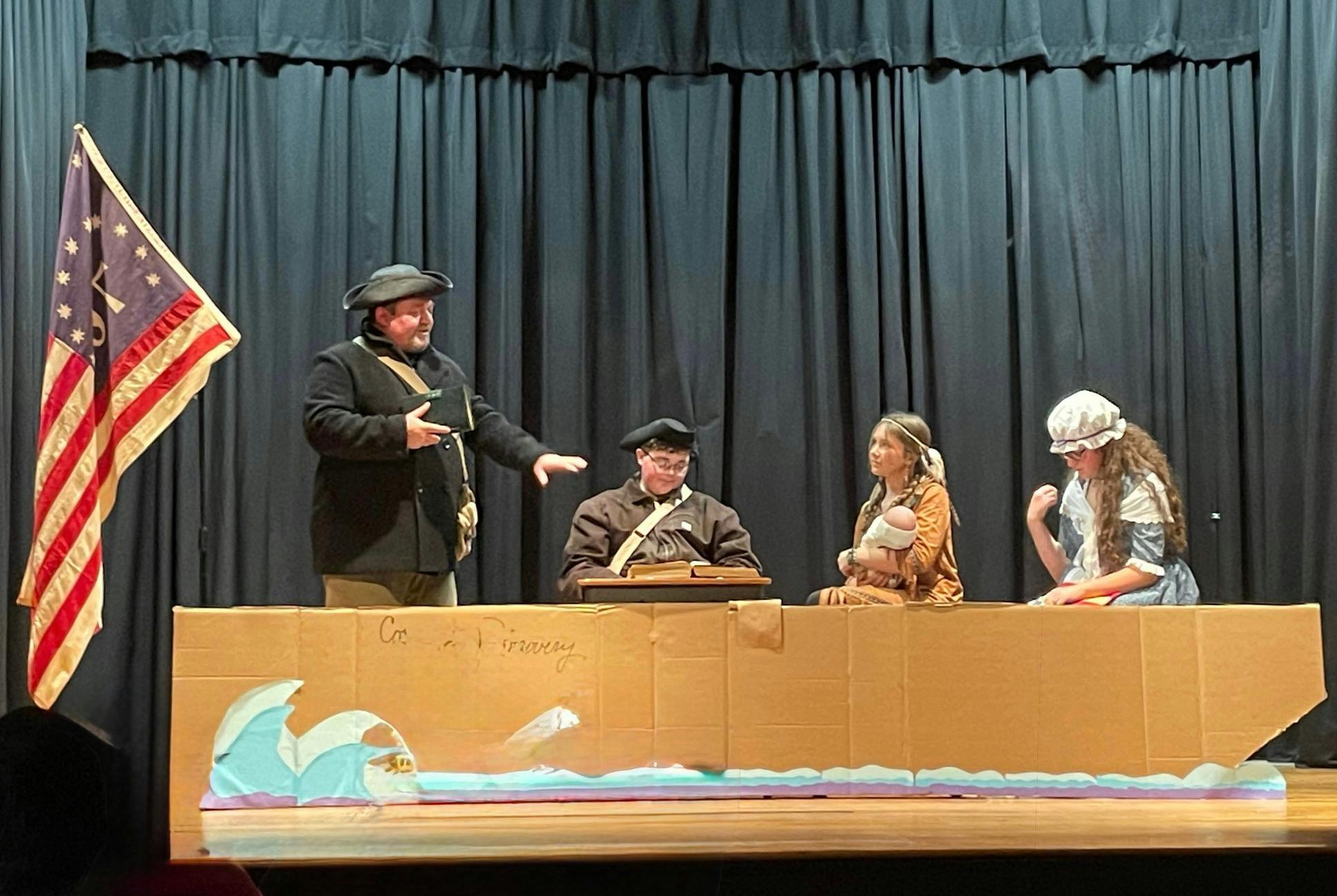 People in period costumes on a stage with a flag and cardboard boat prop