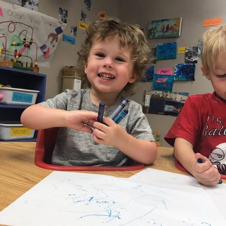 Child holding crayons and smiling while drawing at a table