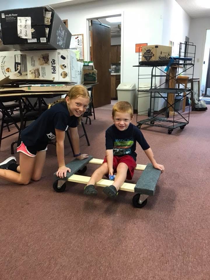 Child kneeling next to another child sitting on a wooden cart with wheels