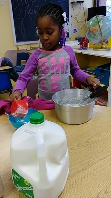 Child holding a red cup over a blue bowl next to a pot and milk jug