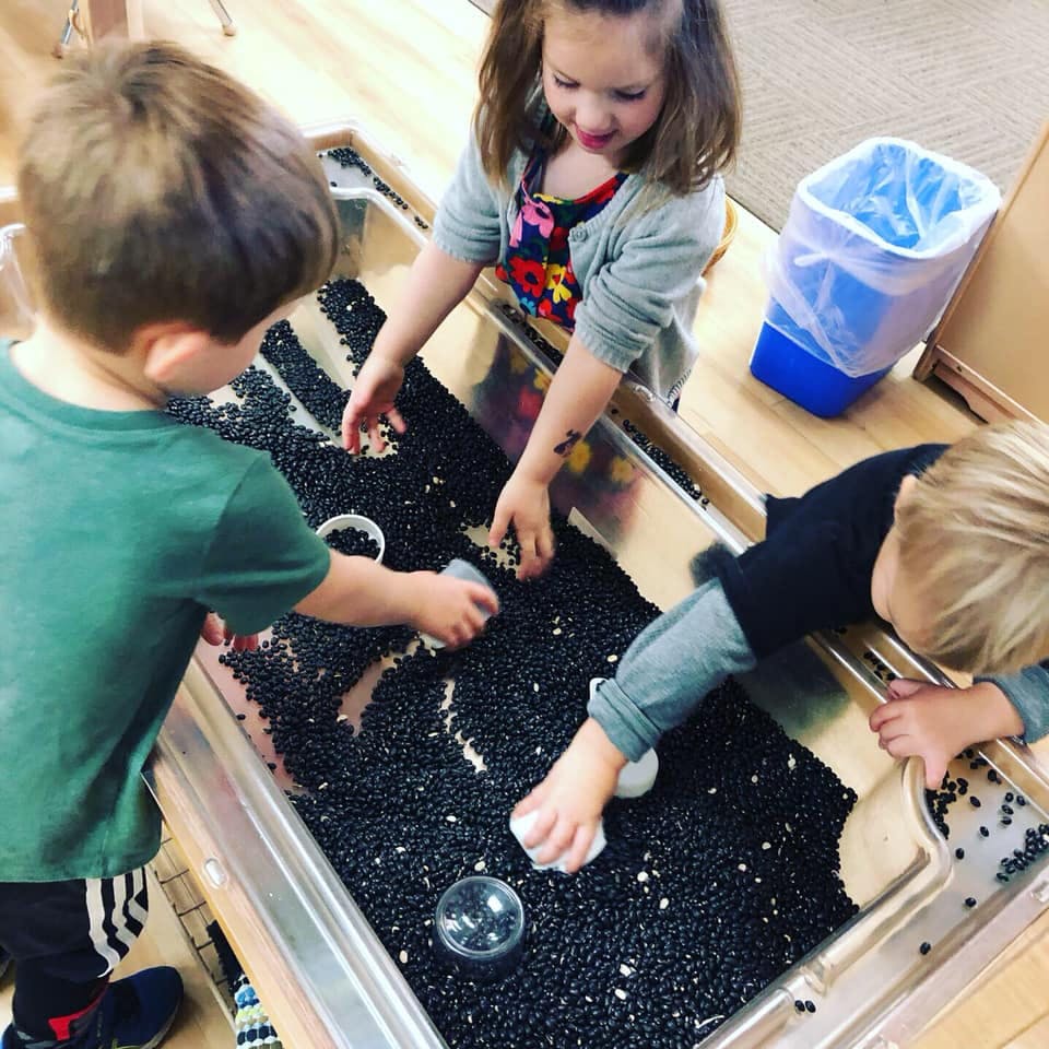 Three children playing with black beans in a sensory bin
