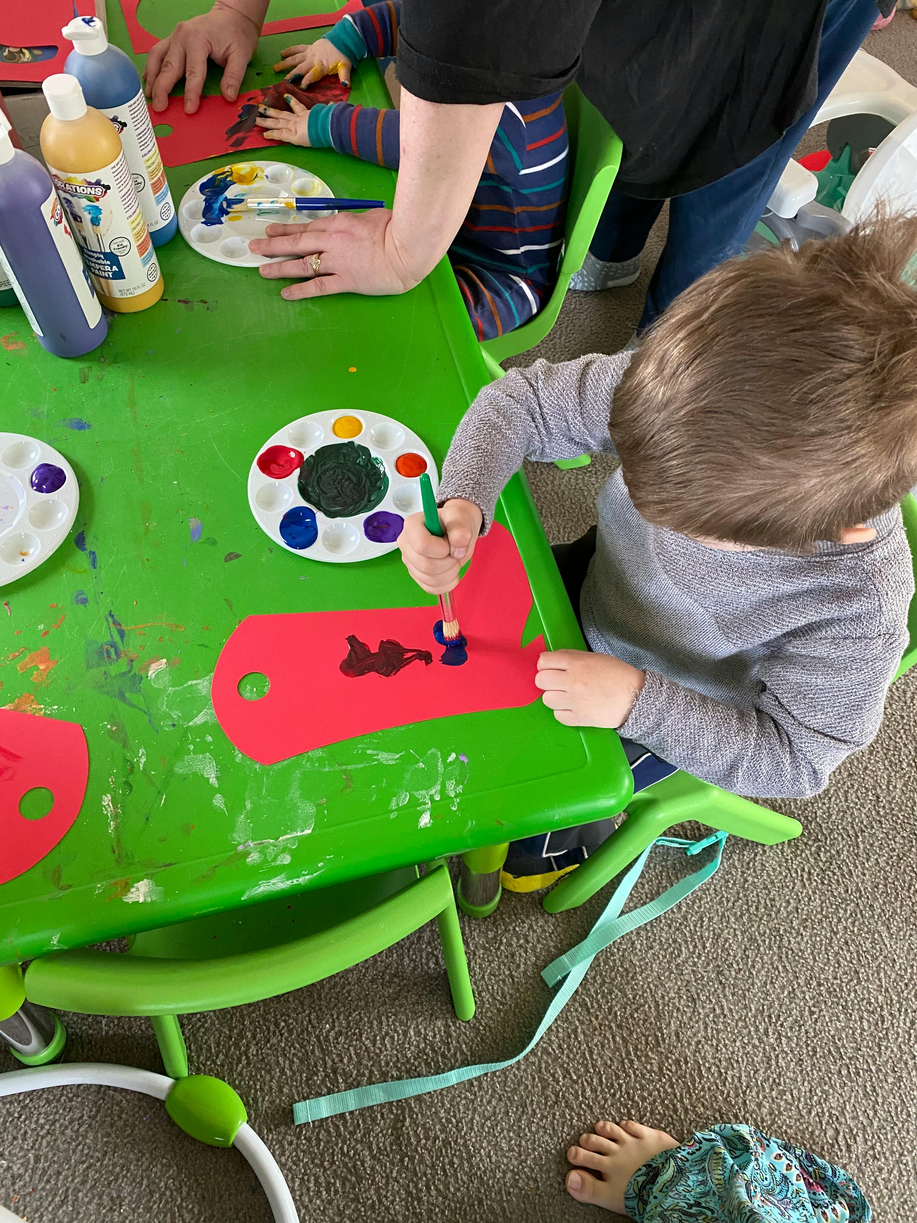 Child painting on red paper at a green table with an adult