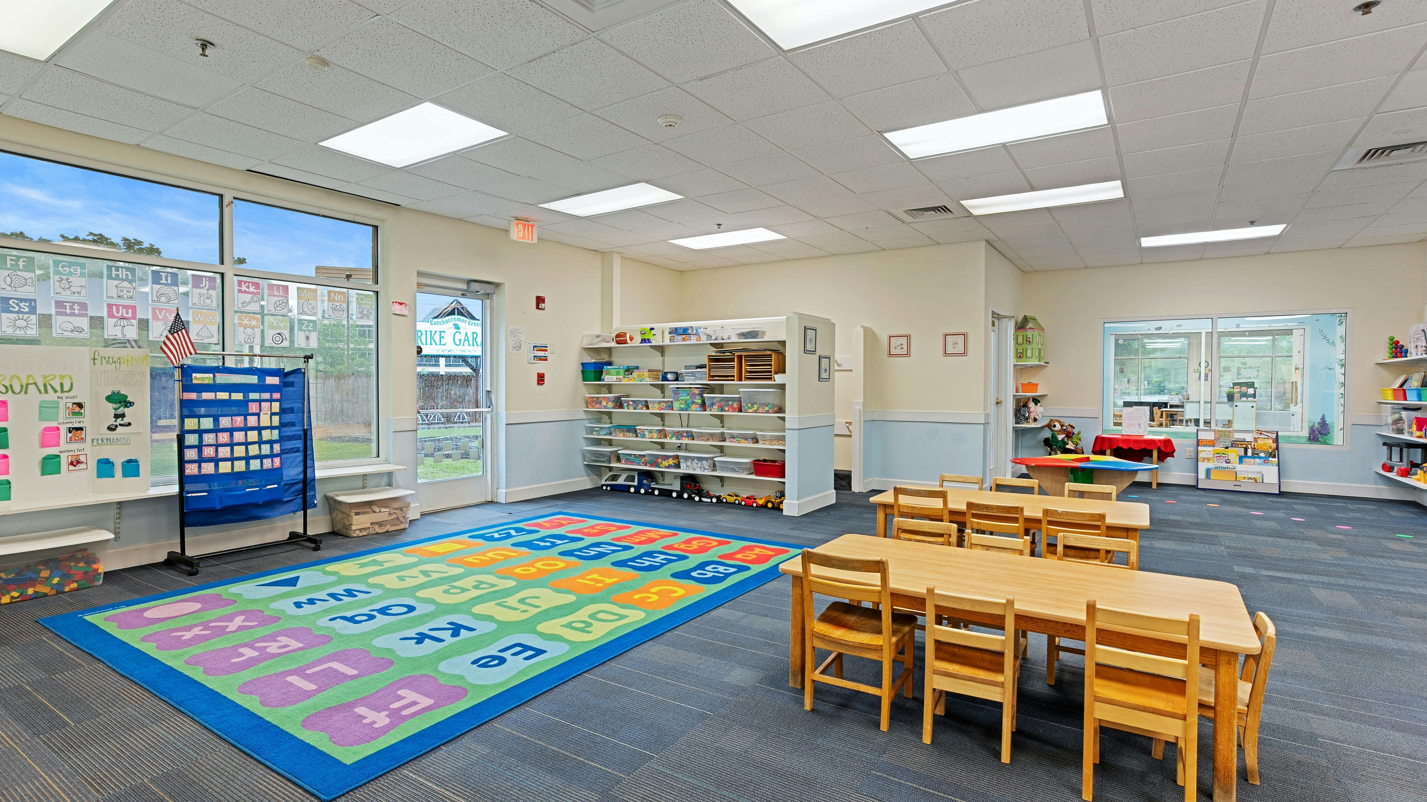 Classroom with child-sized tables and chairs, alphabet rug, and shelves with toys