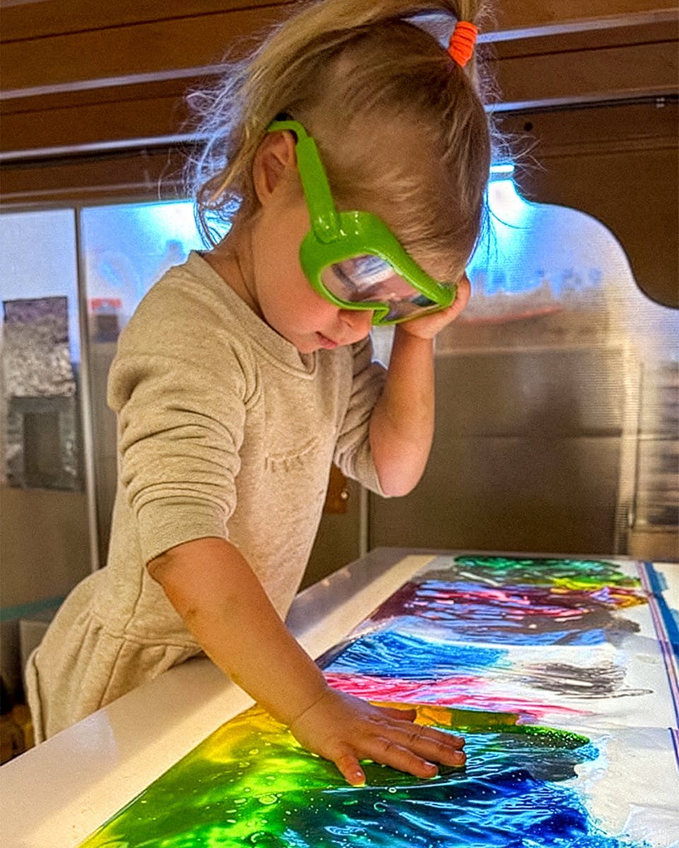 Child wearing green goggles, hand on colorful liquid on a surface