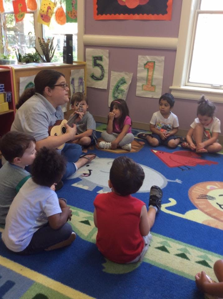 Adult playing guitar for a group of children sitting on a rug