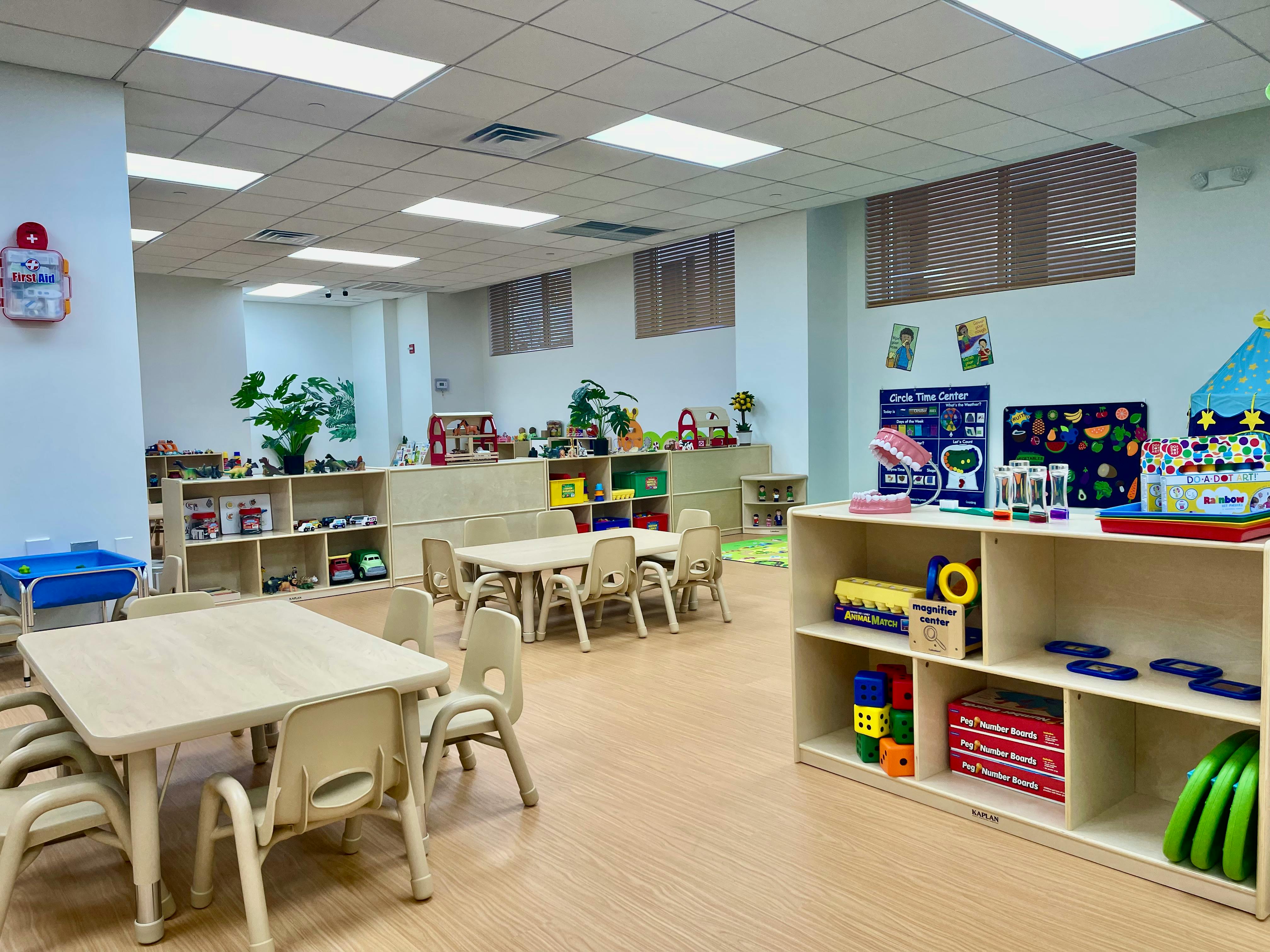 Empty classroom with small tables and chairs, shelves with toys and learning materials