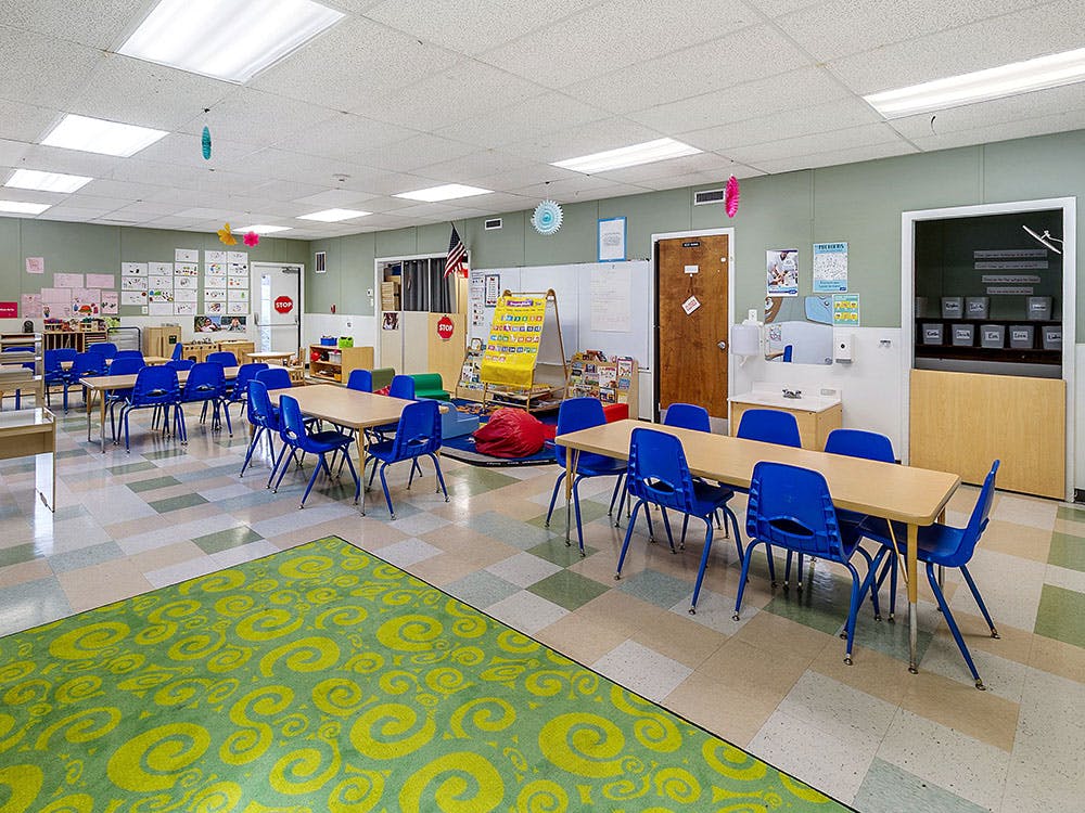 Empty classroom with small tables and blue chairs, green rug with yellow swirls