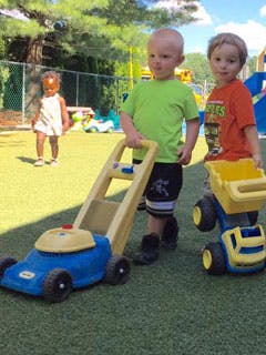 Two children playing with toy lawnmower and dump truck outdoors