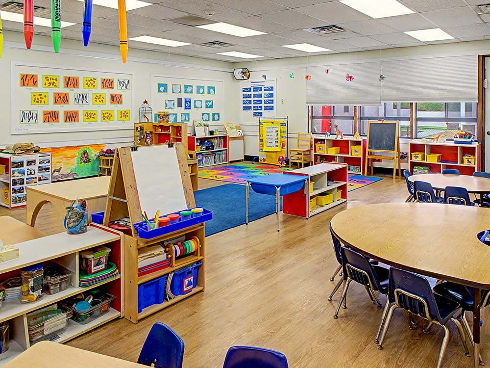 Empty preschool classroom with small tables and chairs, artwork on walls