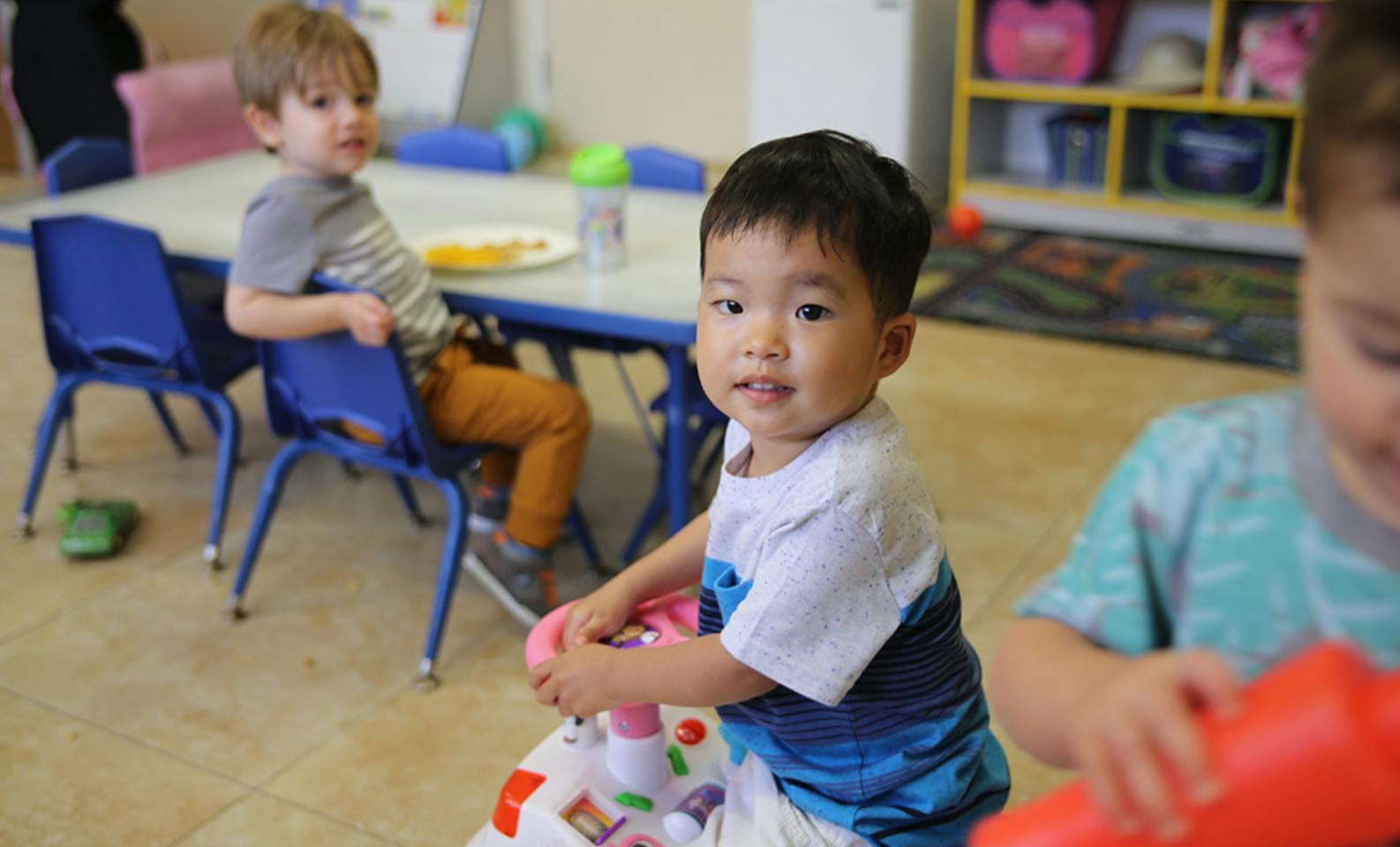 Child playing with toy, another child seated at table in background