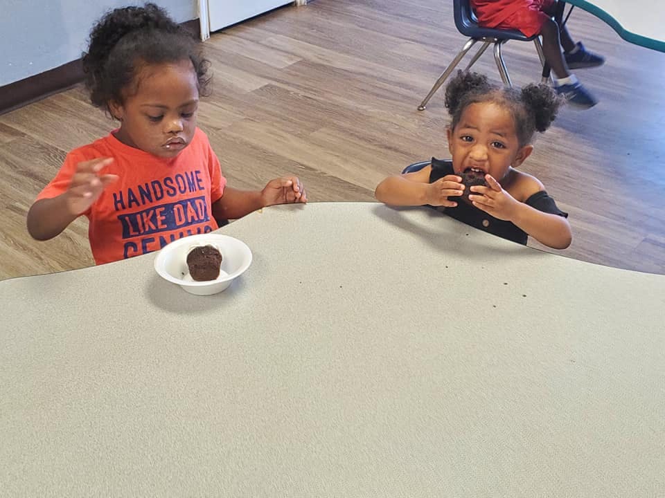 Two children at a table, one eating a cupcake and one looking at a cupcake