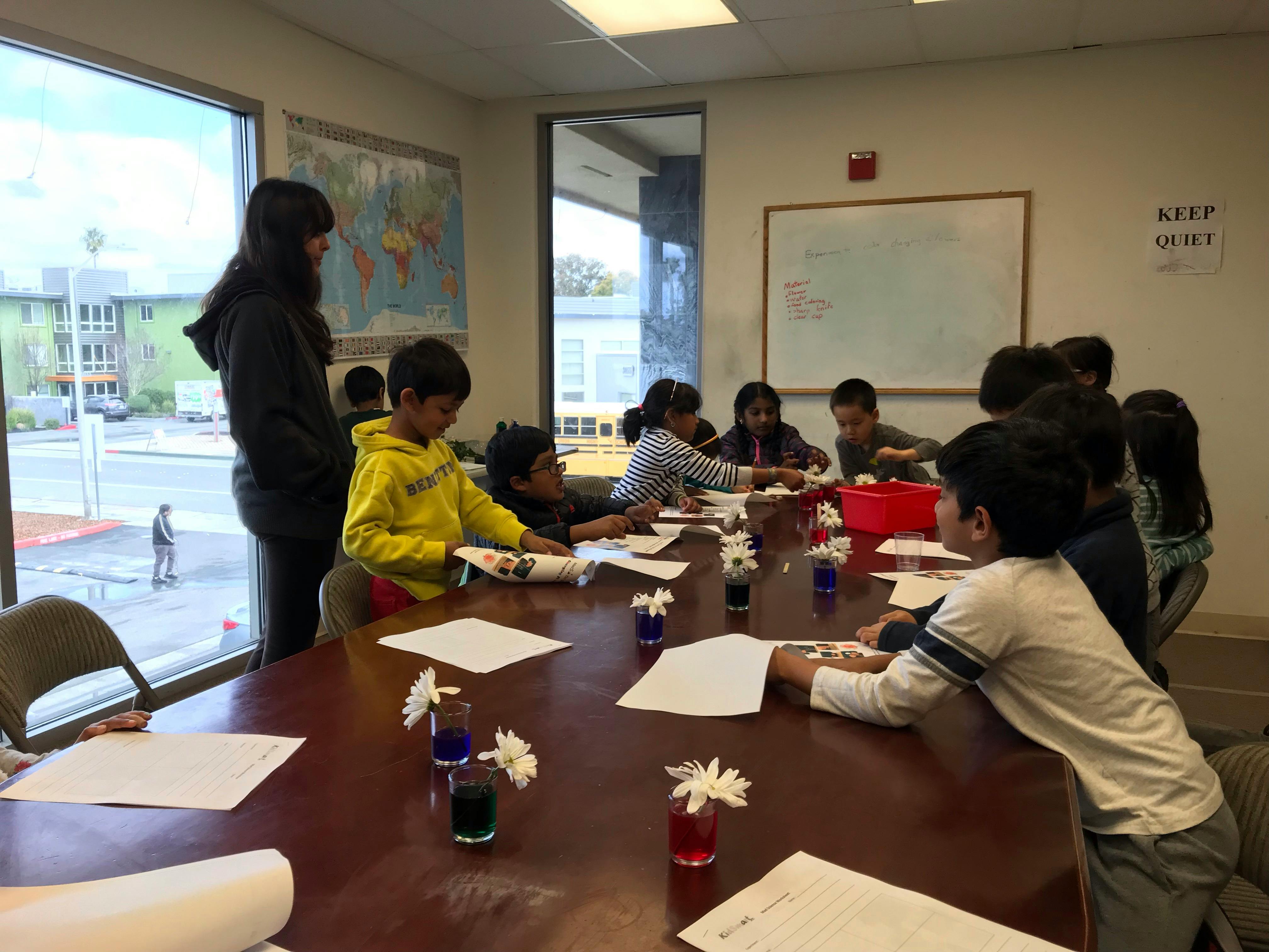 Person standing next to children seated at a table with papers