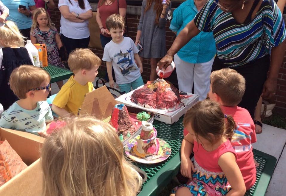 Adult pouring liquid into a volcano model as children watch