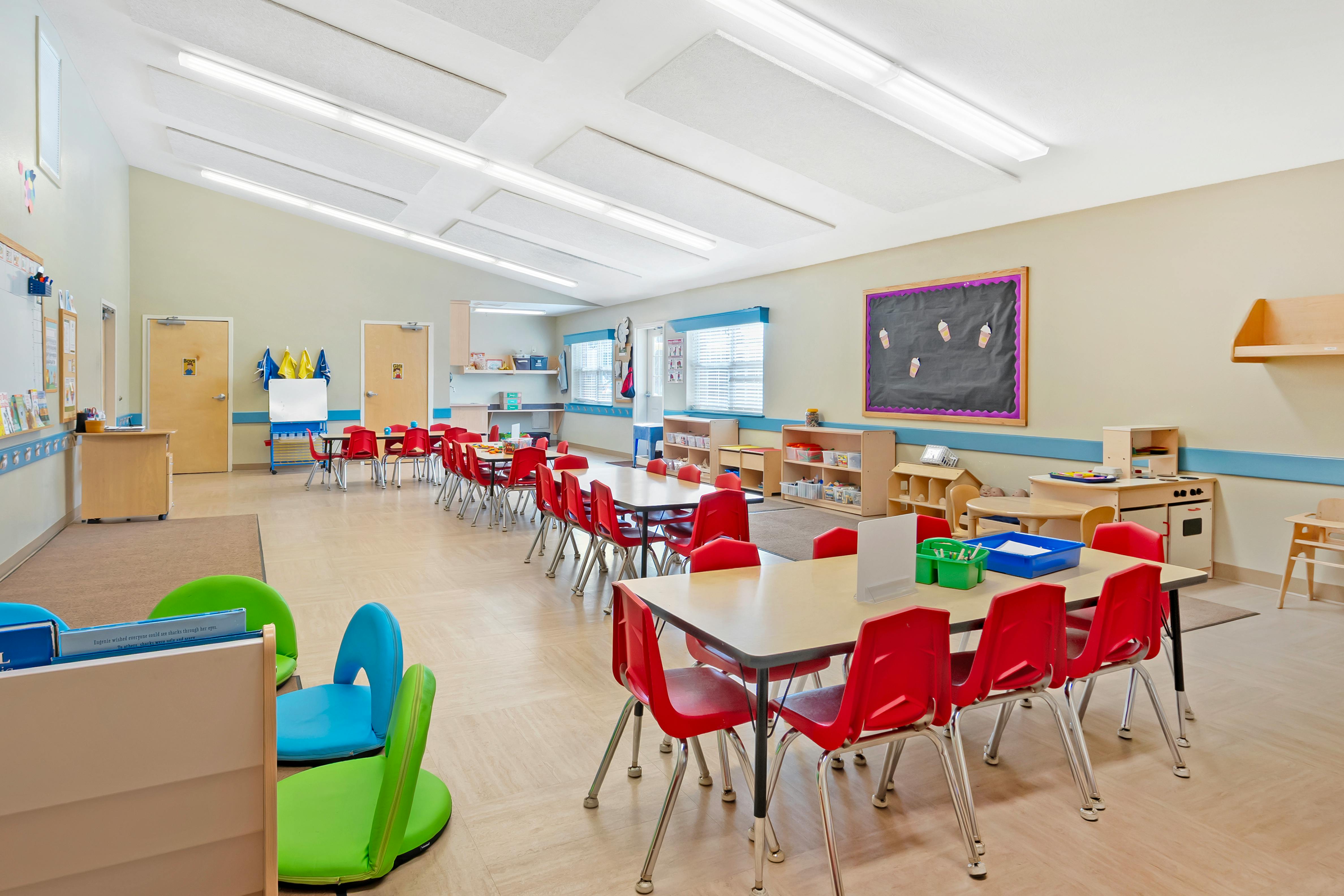 Empty classroom with red chairs and tables, a play kitchen, and a blackboard