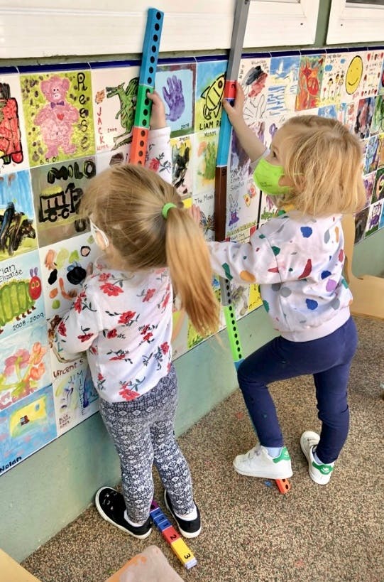 Two children playing with long colorful toys in front of a wall of drawings