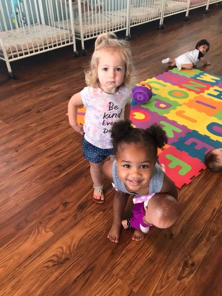 Two children playing on a wooden floor, one holding a doll
