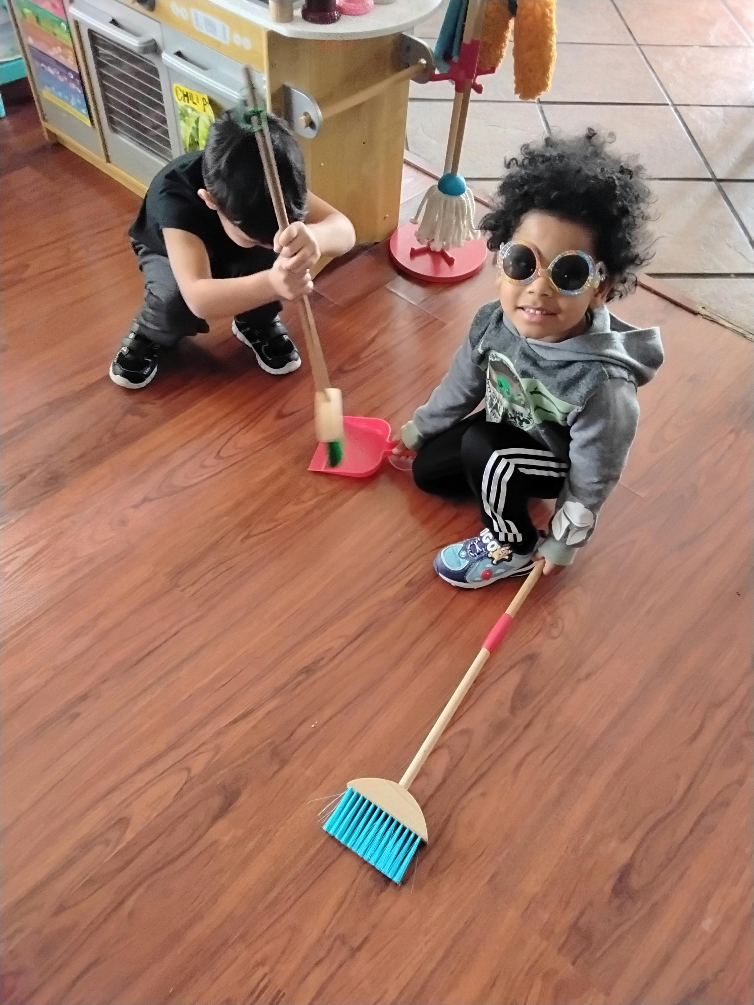 Two children playing with toy cleaning tools on a wooden floor