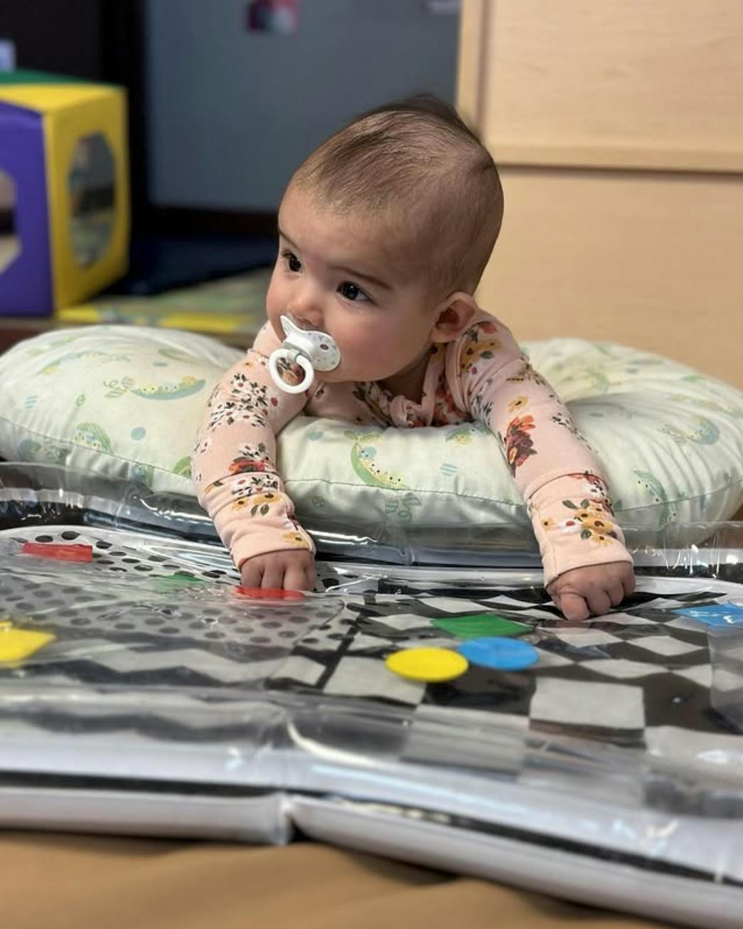 Child with pacifier doing tummy time on a patterned mat with colorful shapes