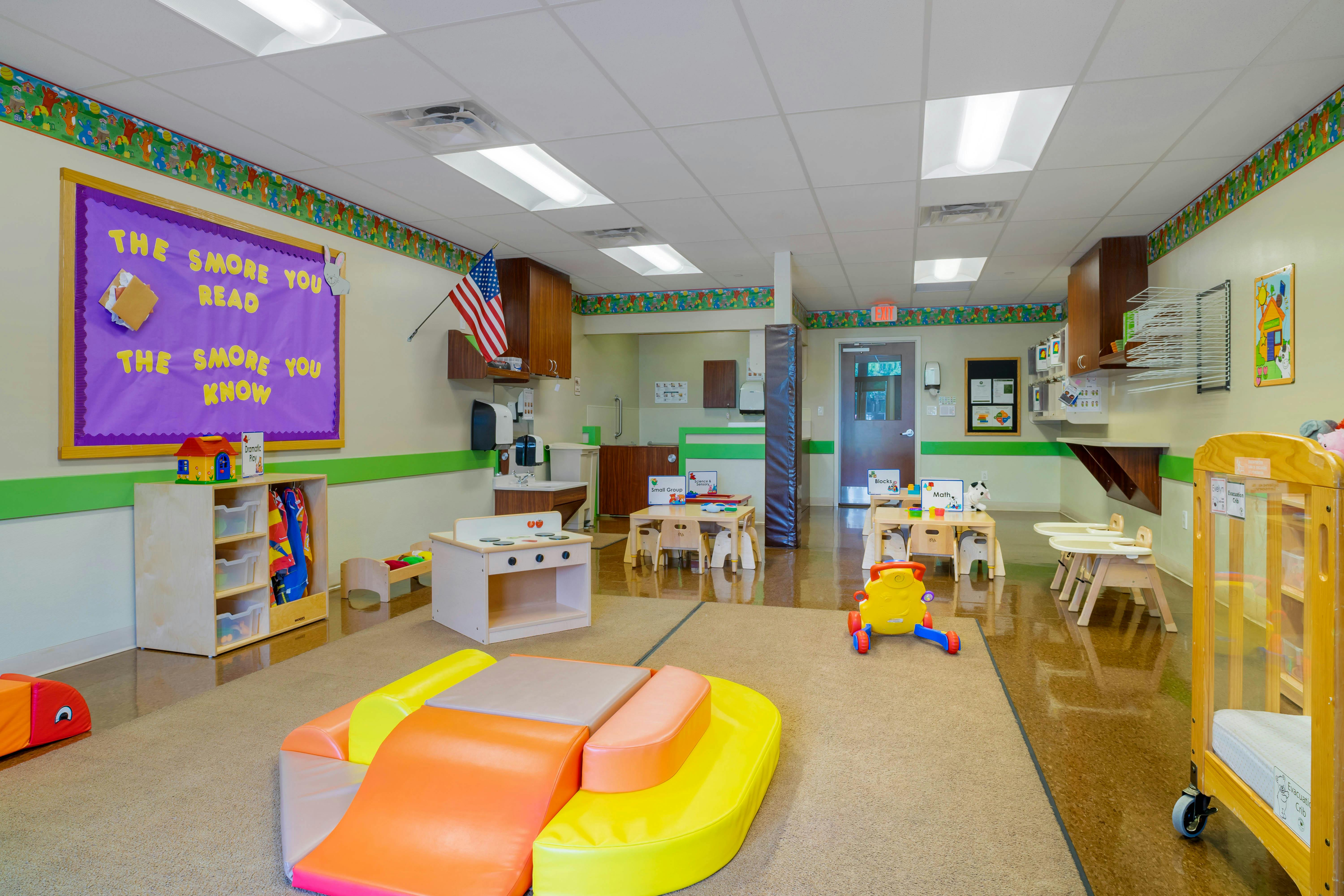 A colorful empty classroom with a bulletin board, play kitchen, and activity tables