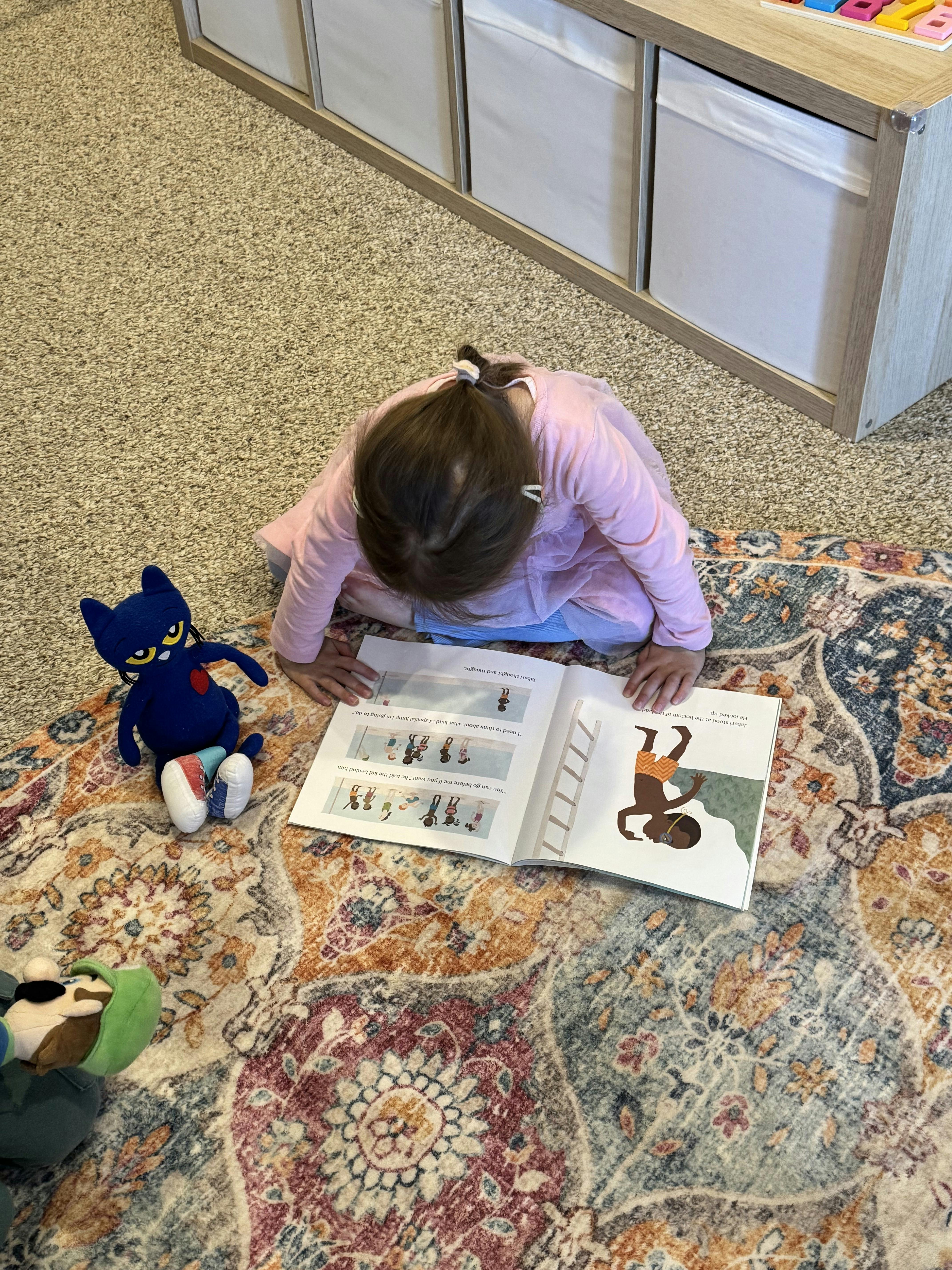 Child on a rug looking at an open book with a blue stuffed animal nearby