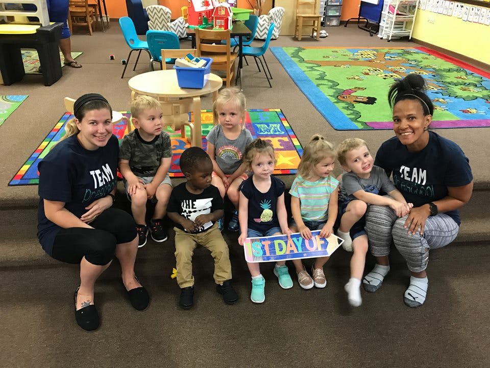 Two people and several children sitting in a classroom, one child holding a "1st Day Of" sign