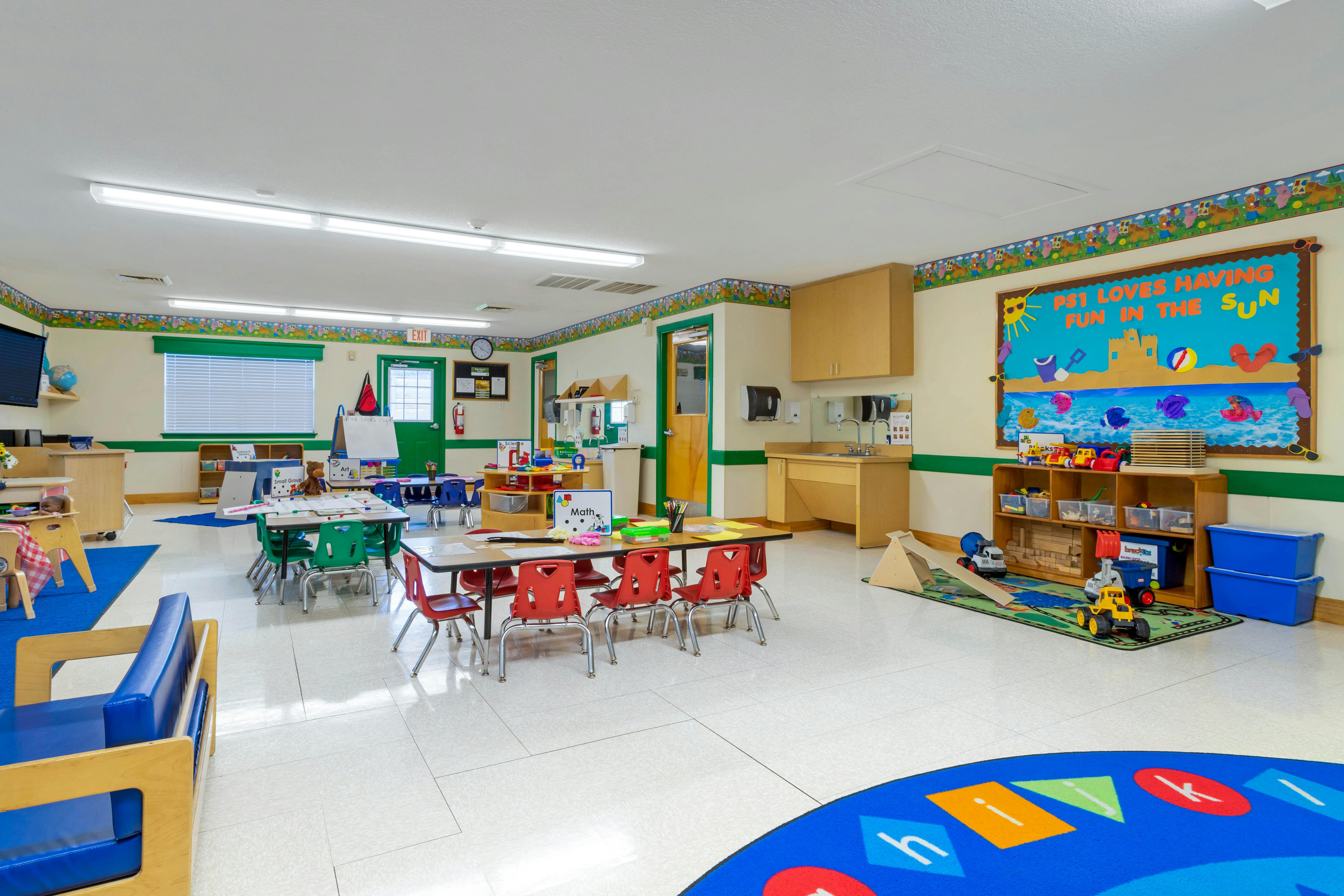An empty preschool classroom with small tables and chairs, toys, and a sink area