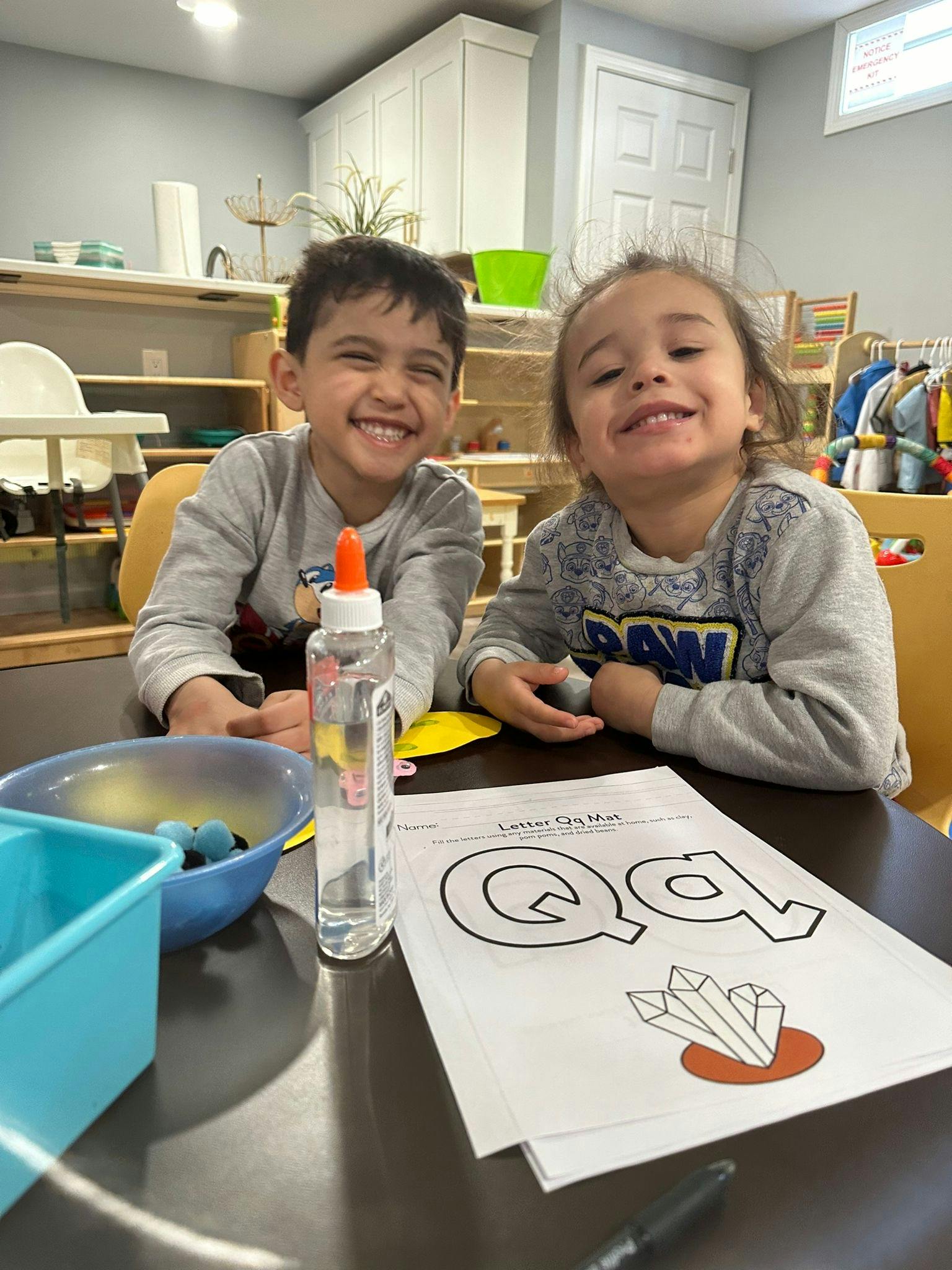 Two children sitting at a table with a letter Q worksheet and glue