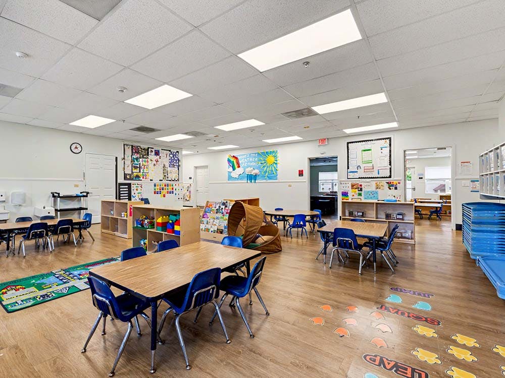 Empty classroom with small tables and chairs, shelves with toys, and wall decorations