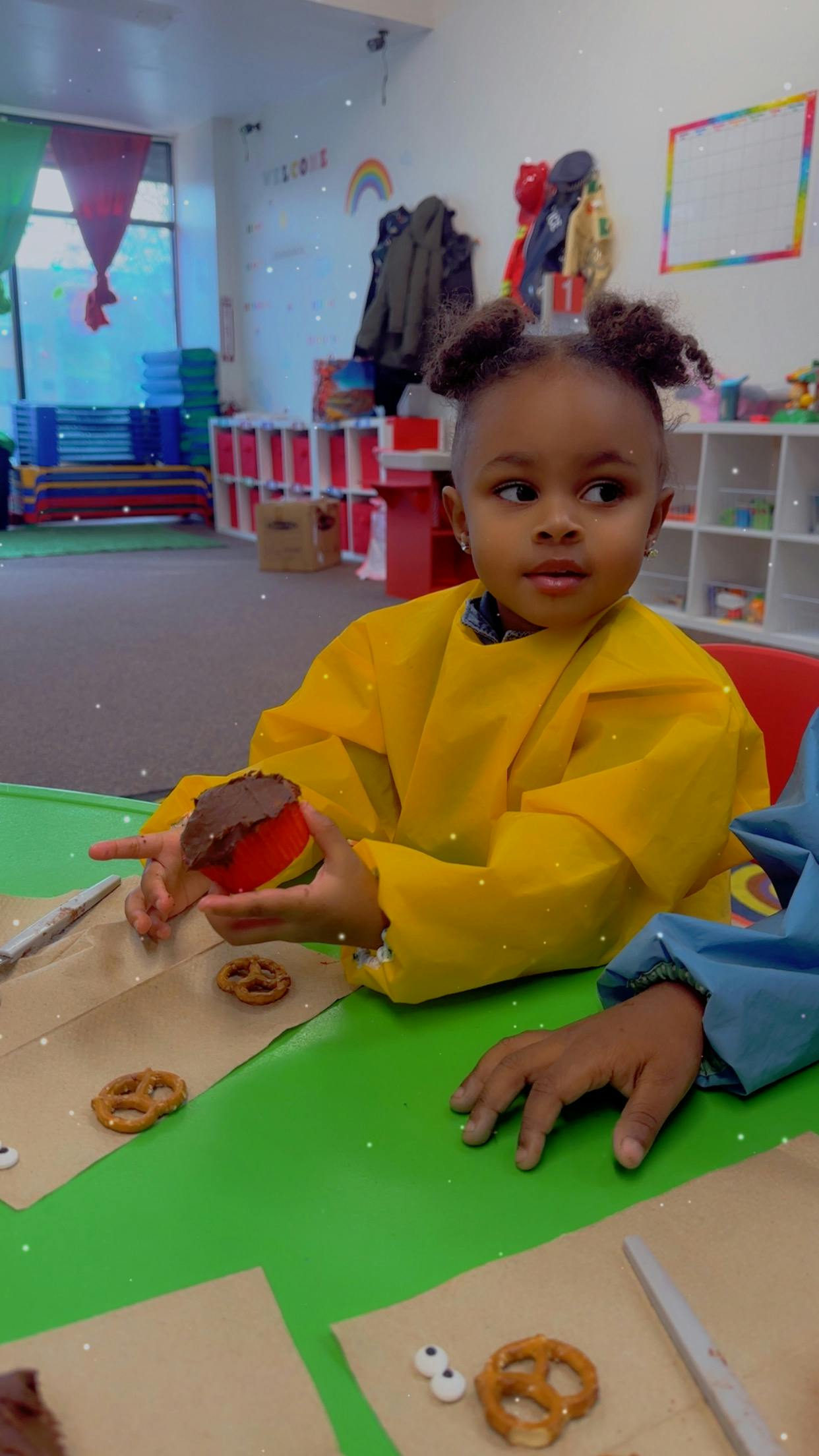 Child in yellow smock holding a cupcake in a classroom