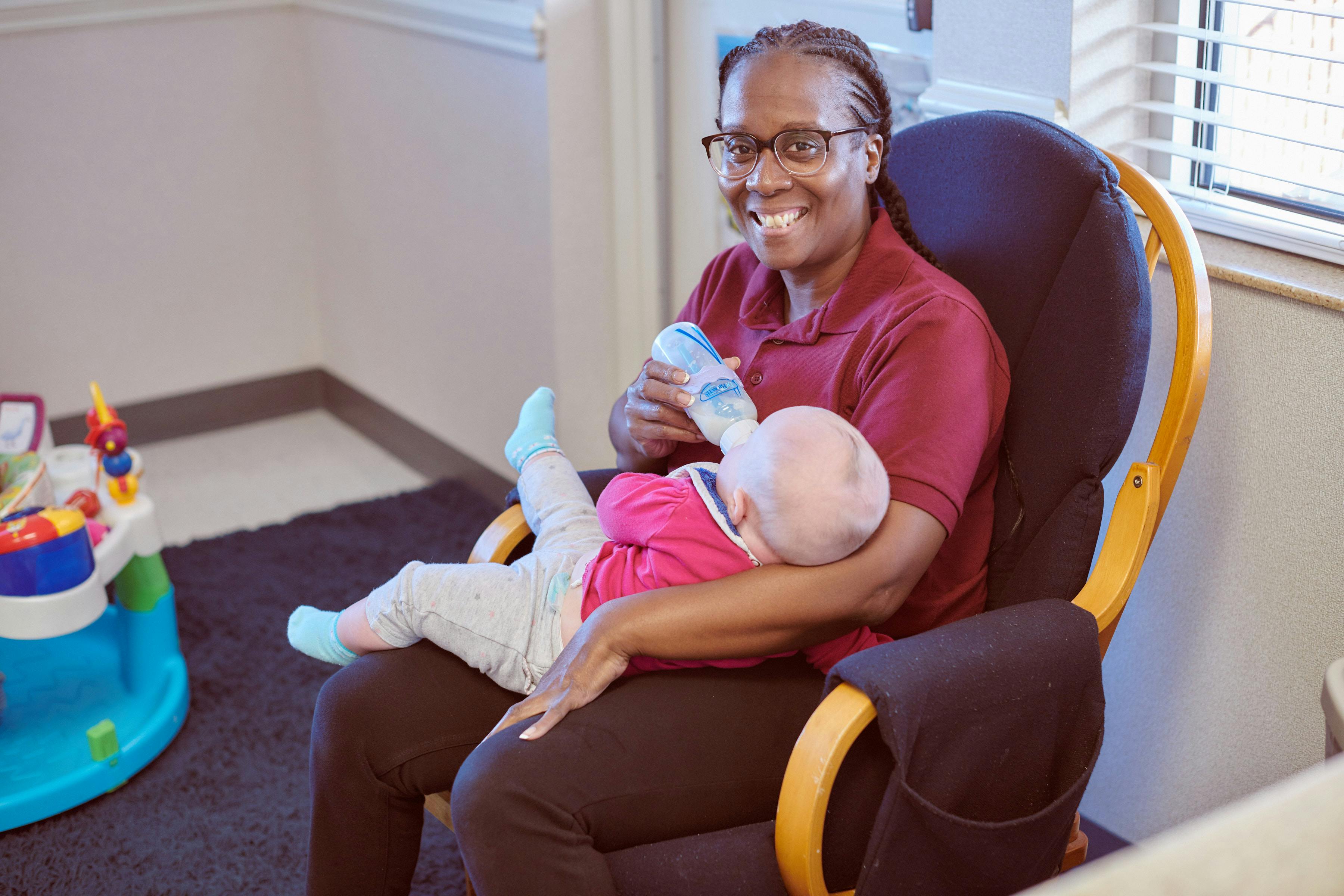 Person in glasses feeding a baby a bottle while sitting in a chair