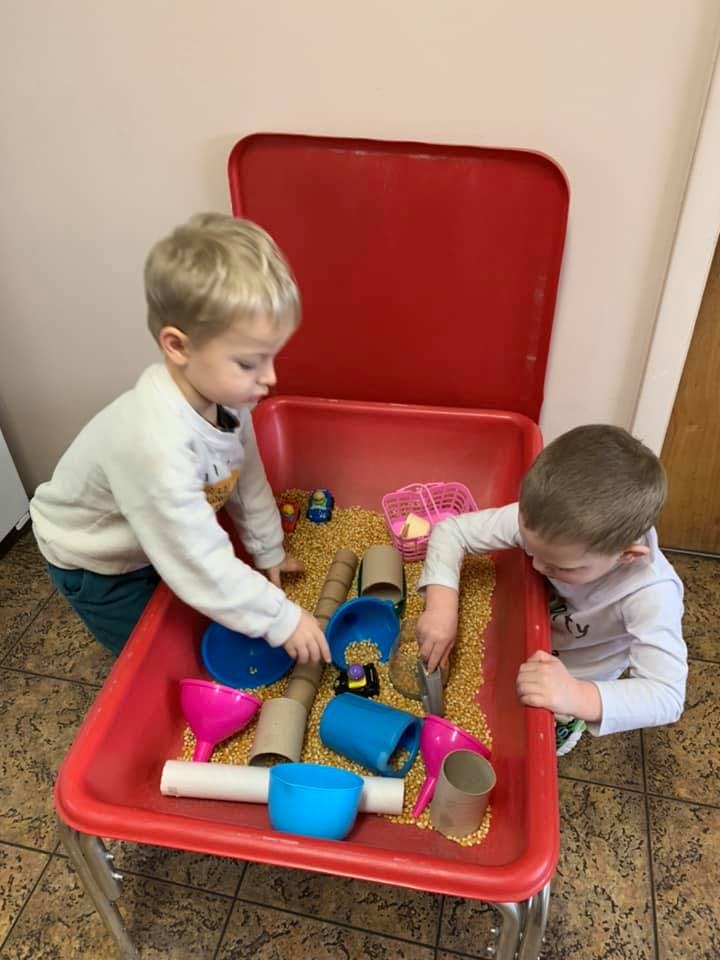 Two children playing with corn kernels and various objects in a red bin