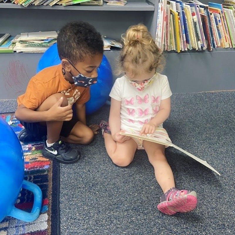 Two children wearing masks looking at a book on the floor
