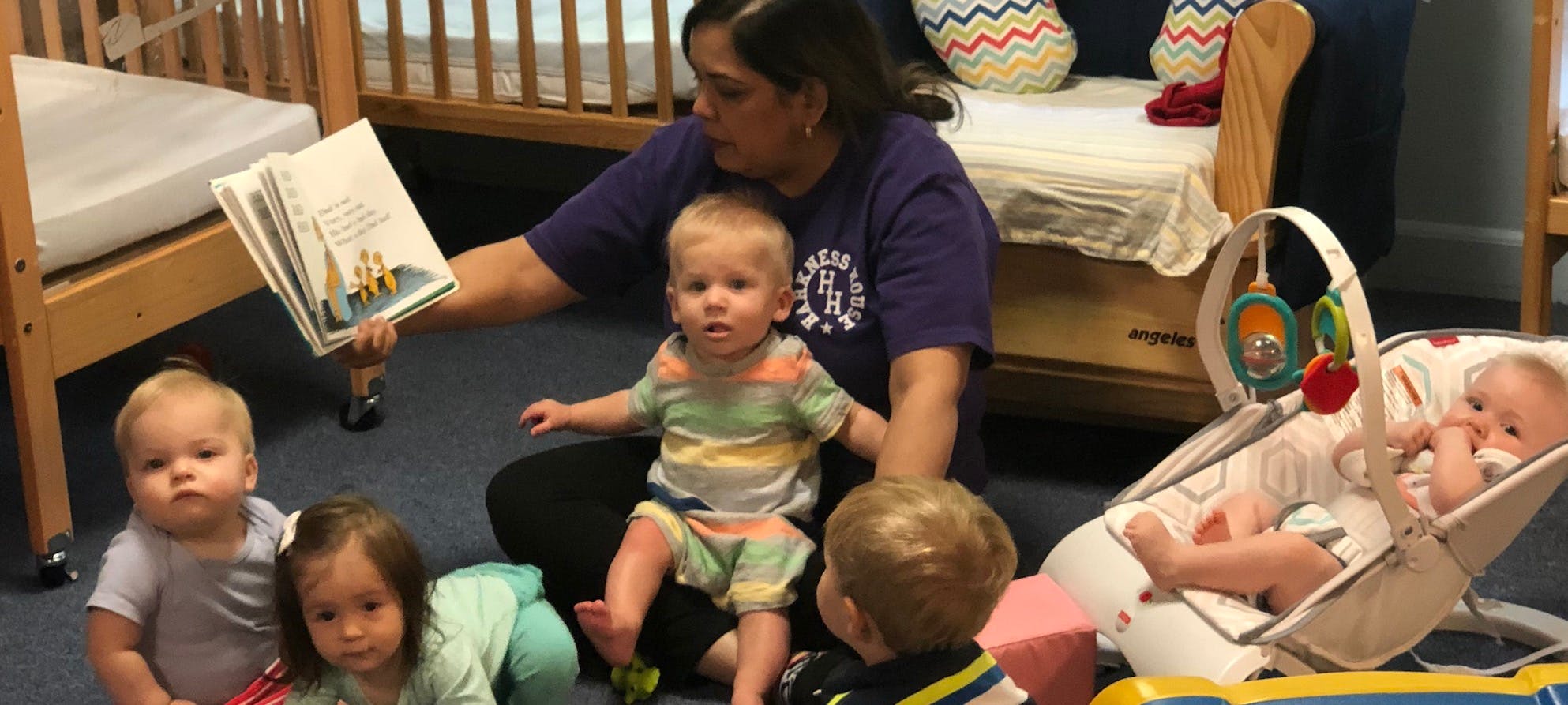 Person reading a book to multiple children in a room with cribs in the background