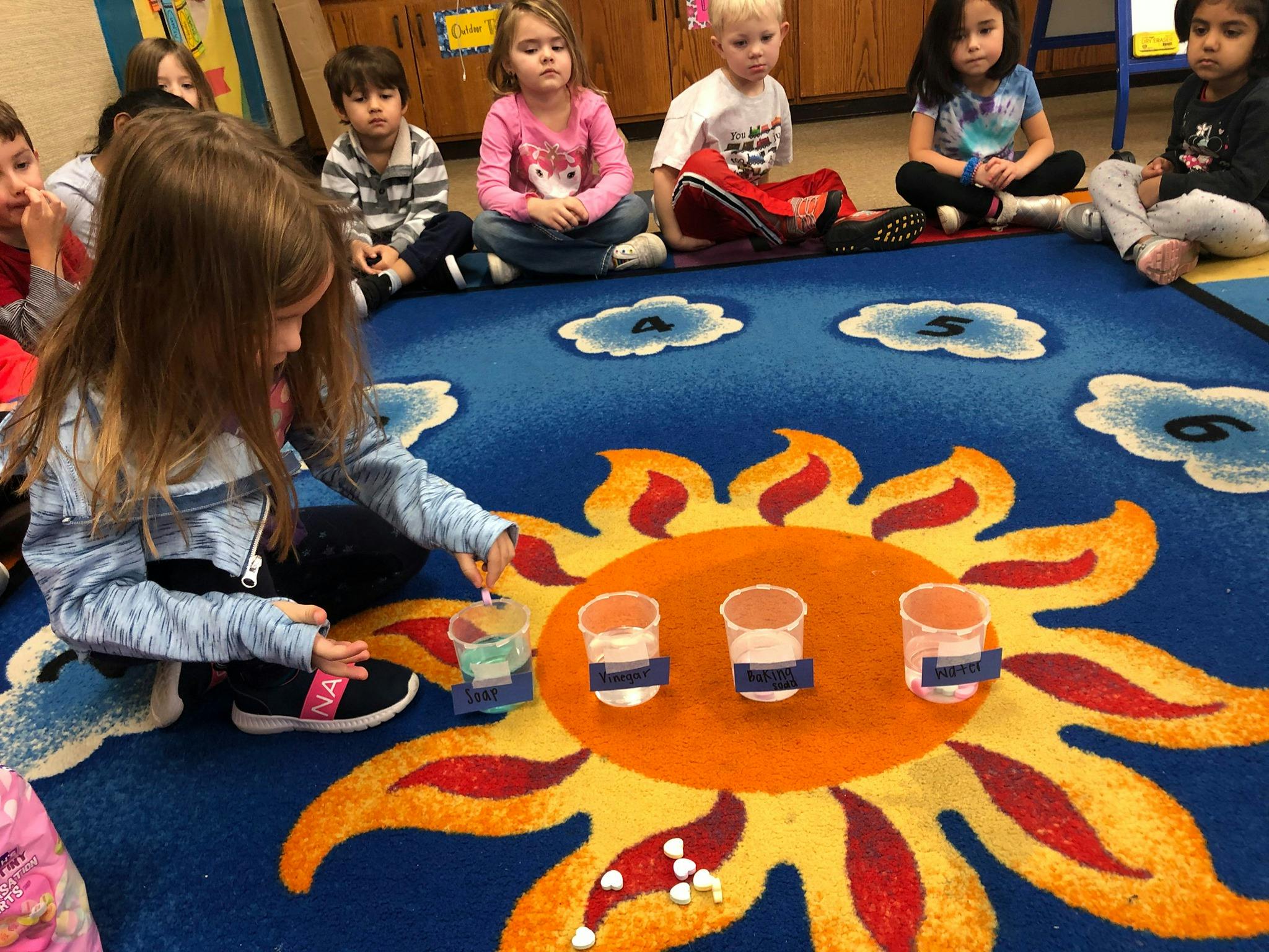 Child playing with cups on a sun-patterned rug with other children watching
