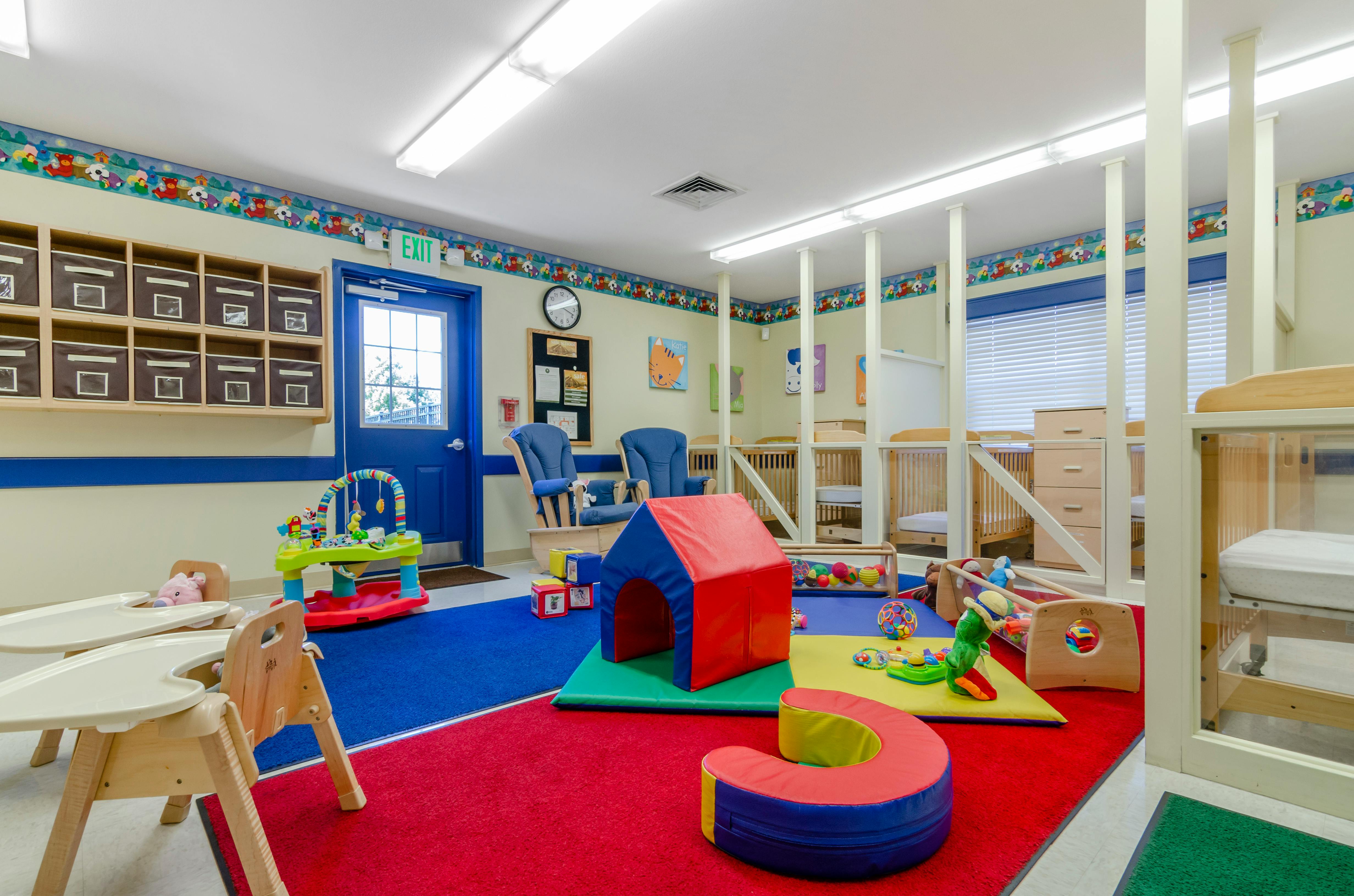 A childcare room with colorful rugs, cribs, high chairs, and play equipment