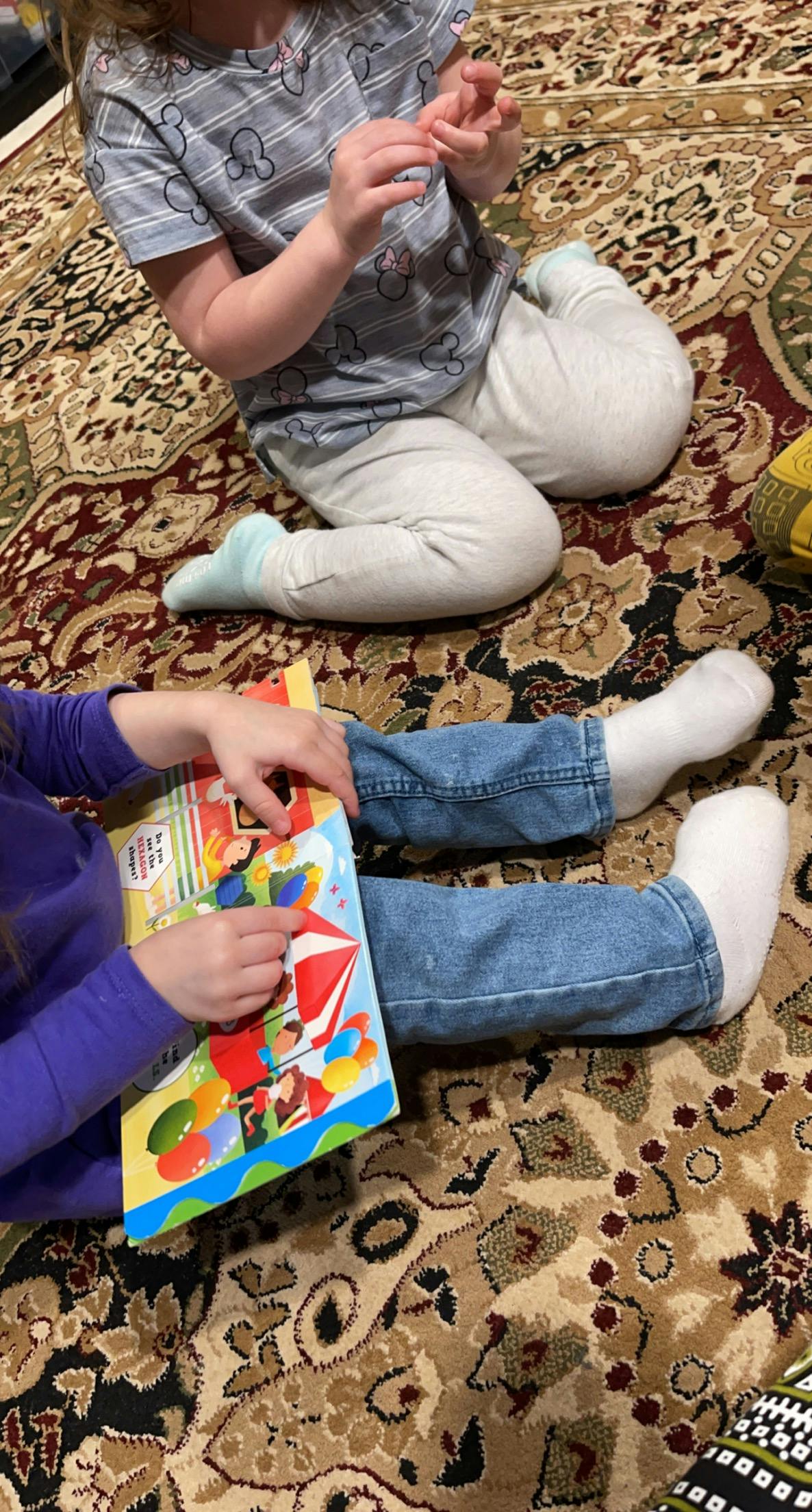 Two children sitting on a patterned rug, one holding a colorful book