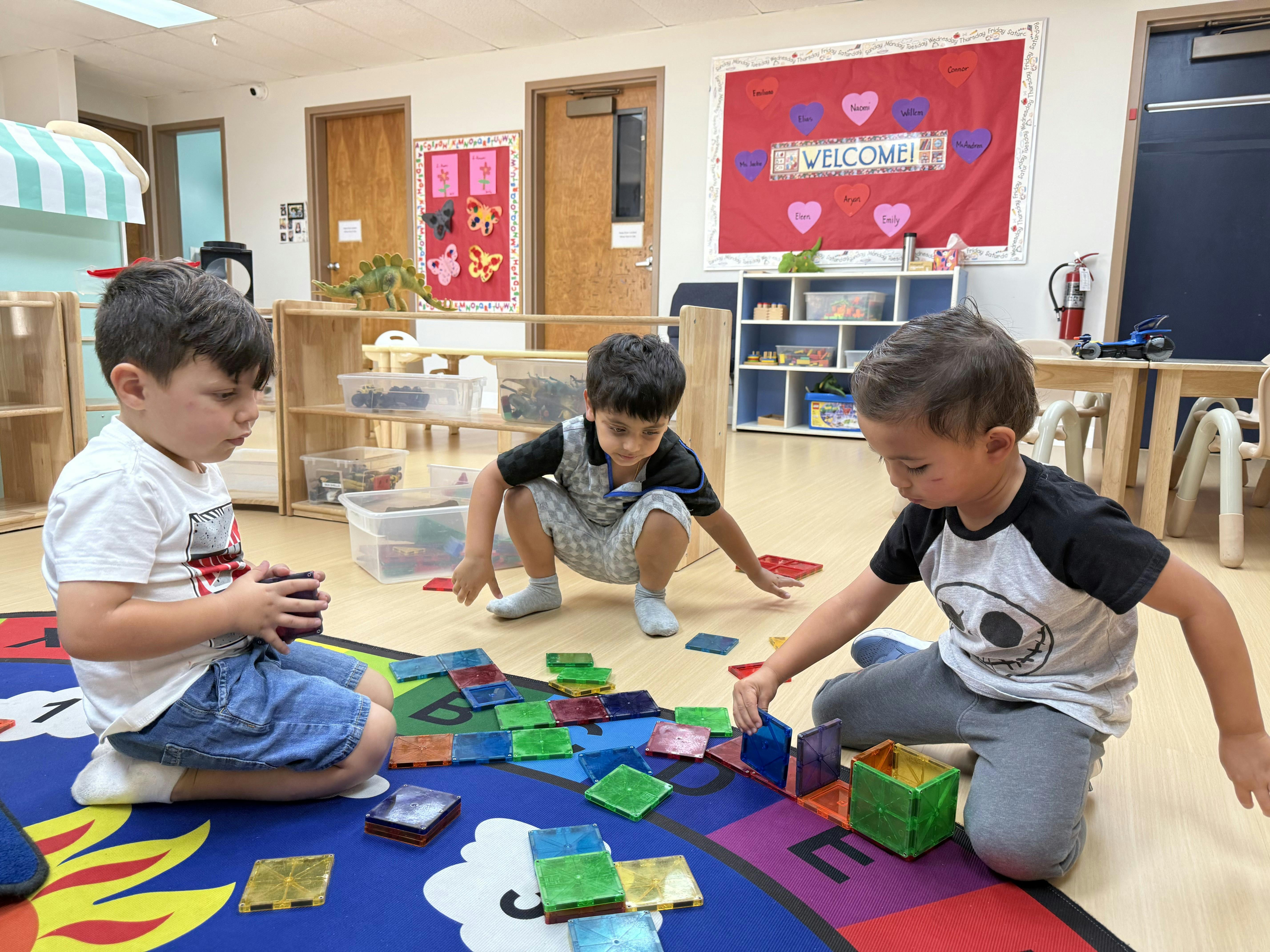 Three children playing with colorful magnetic tiles on a rug in a classroom