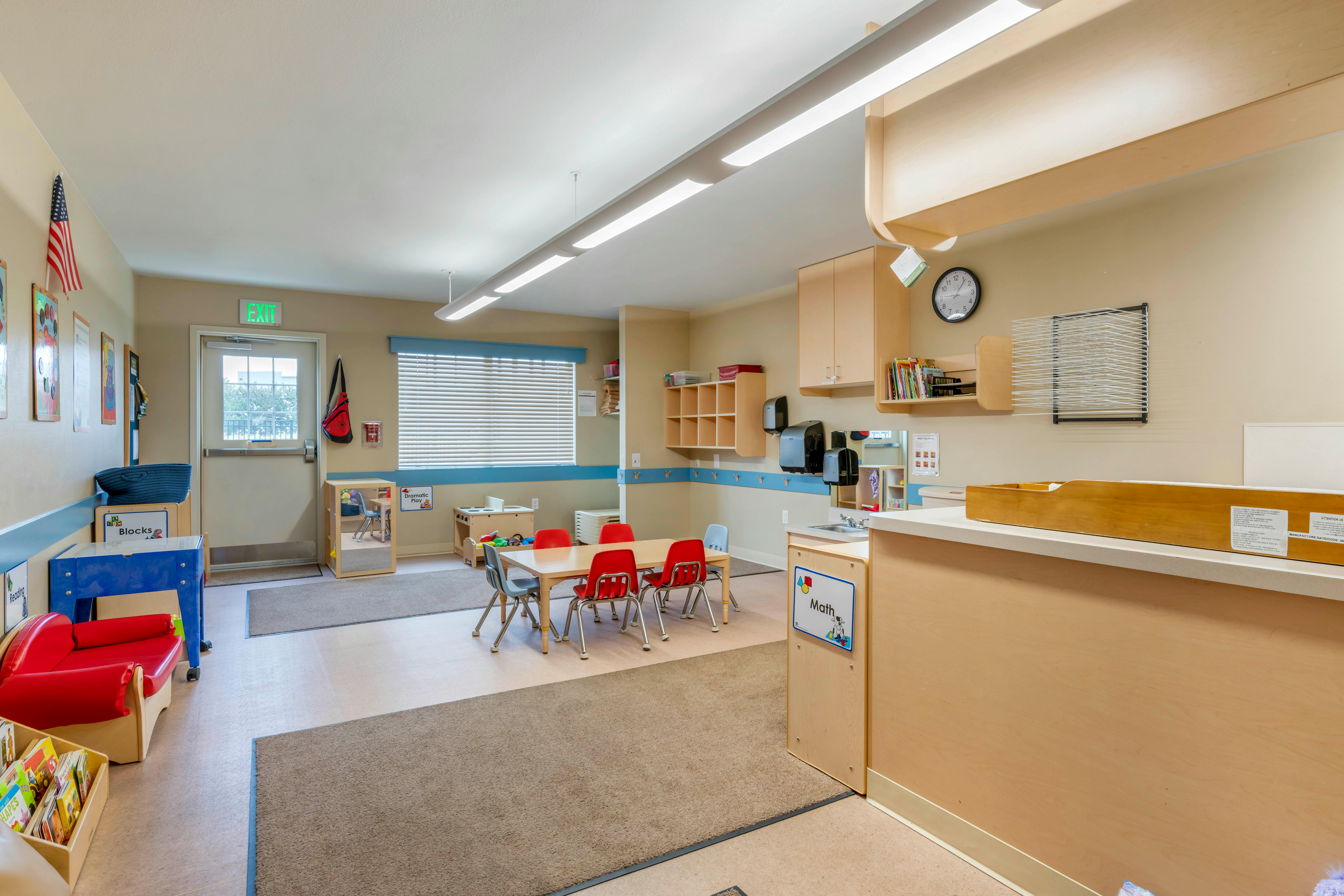 A brightly lit classroom with a table, chairs, play area, and an exit door