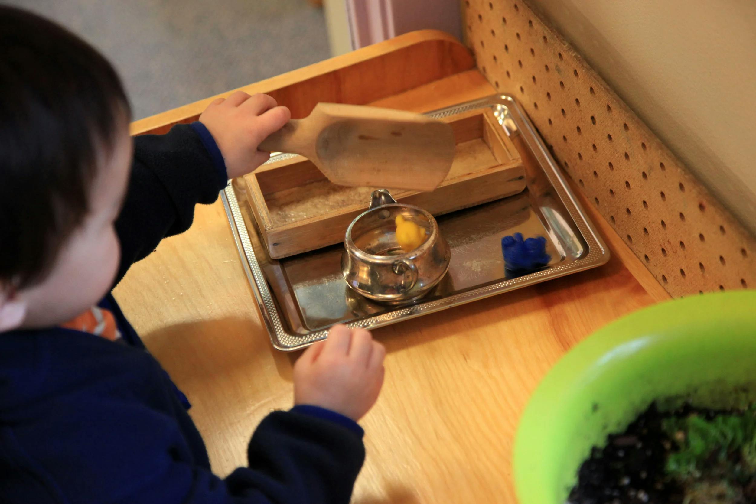 Child's hands playing with a wooden scoop and metal containers on a tray