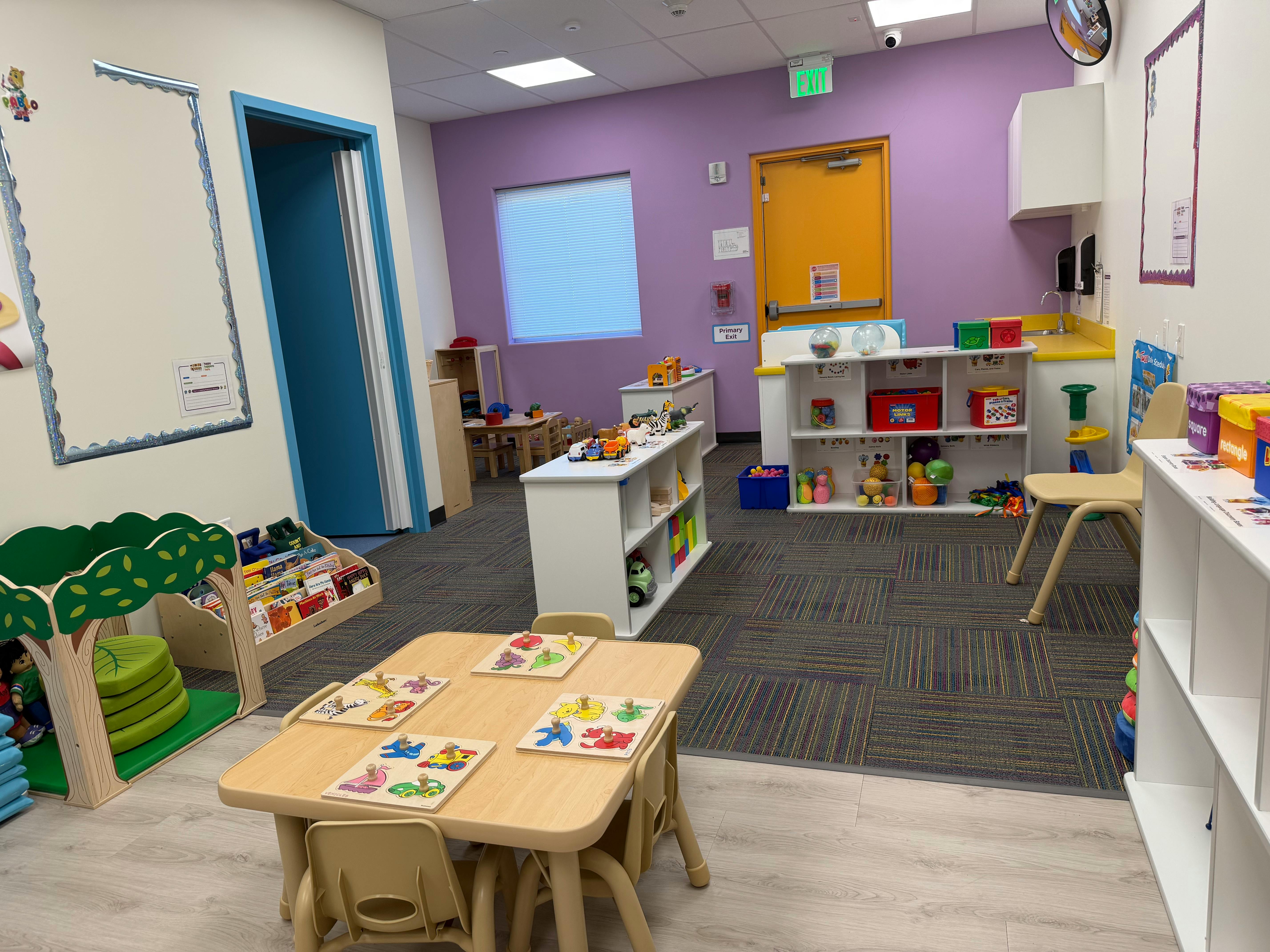 Children's classroom with small tables, shelves of toys, and a primary exit door