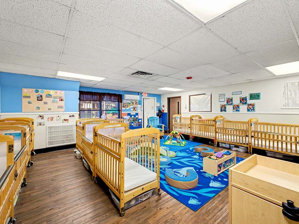 Rows of wooden cribs in a daycare room with a blue play rug