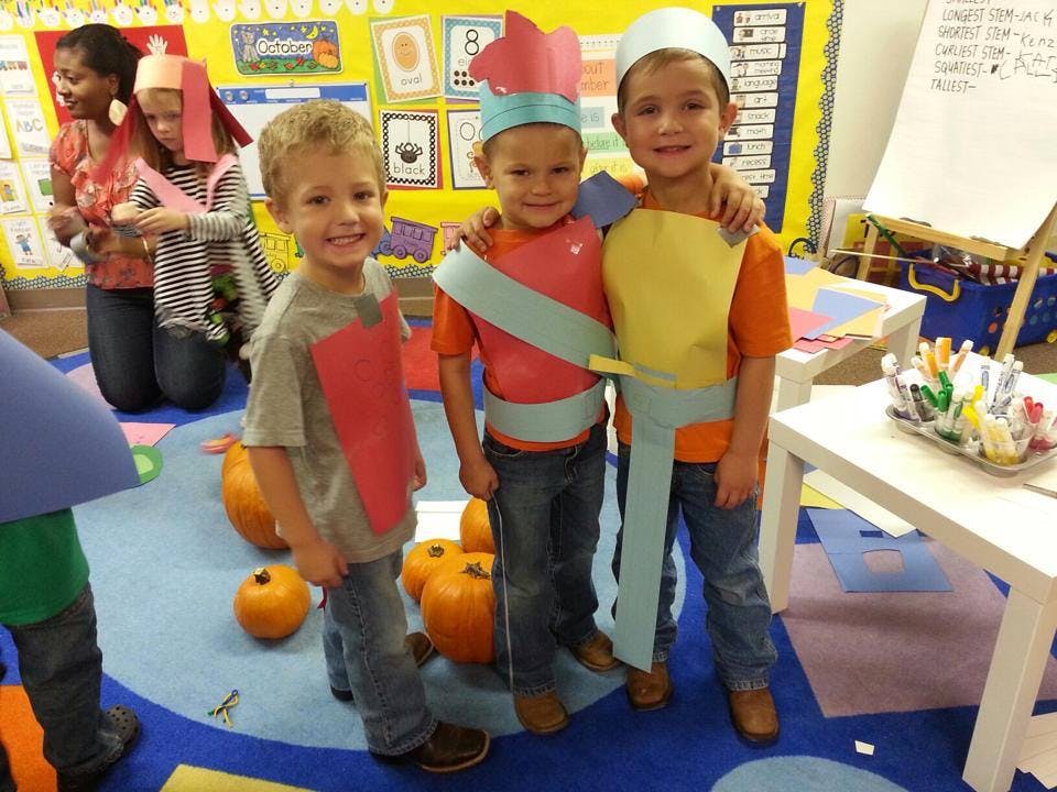 Three children in costumes standing in a classroom with pumpkins on the floor